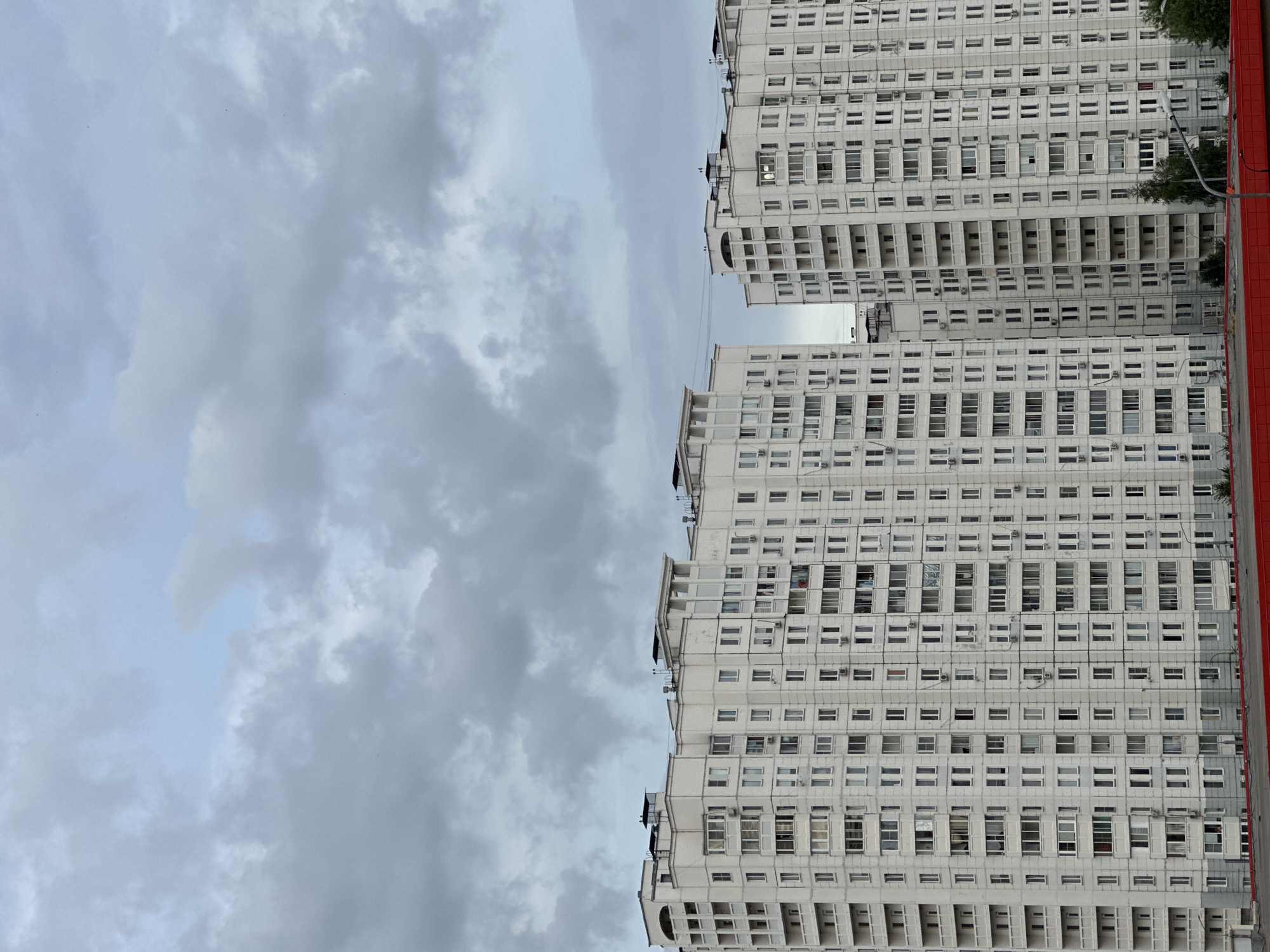 The image shows a view of two residential apartment buildings in Podolsk, Russia, under a cloudy sky. The buildings are tall, with many floors and windows, and are constructed in a typical Soviet-era style. They are primarily white with some gray accents. The sky is overcast with light gray clouds. In the foreground, there is a red structure, possibly a fence or rooftop, that spans across the bottom of the image. There are no people visible in the image. The buildings are located in a densely populated urban area.