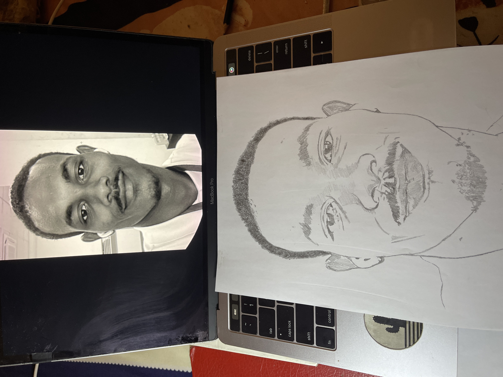 The image shows a partially completed pencil drawing of a man's face resting on an open silver MacBook Pro laptop. On the laptop screen is a grayscale photo of the same man, turned sideways. The keyboard of the laptop is visible beneath the drawing. Part of a sticker is visible on the body of the laptop. The environment is a somewhat cluttered surface with other objects partially visible. There is no information to suggest the location is in Jos, Nigeria.