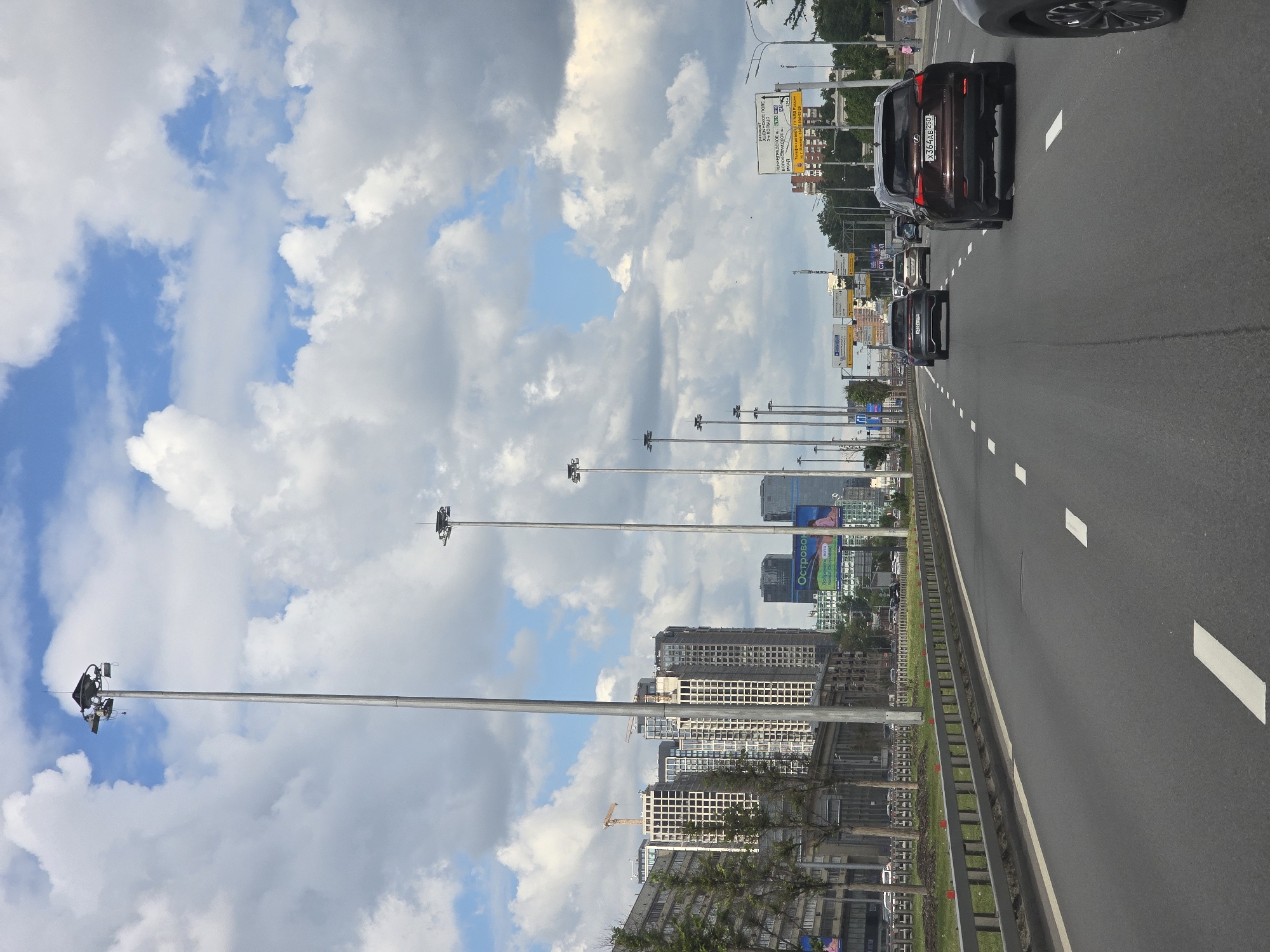 A highway in Moscow, Russia, is shown under a cloudy sky. The road is lined with tall street lights and a barrier fence. Cars are on the right side of the road and move away from the viewer. Buildings are also shown on the right side. A blue and white sky is on the left.