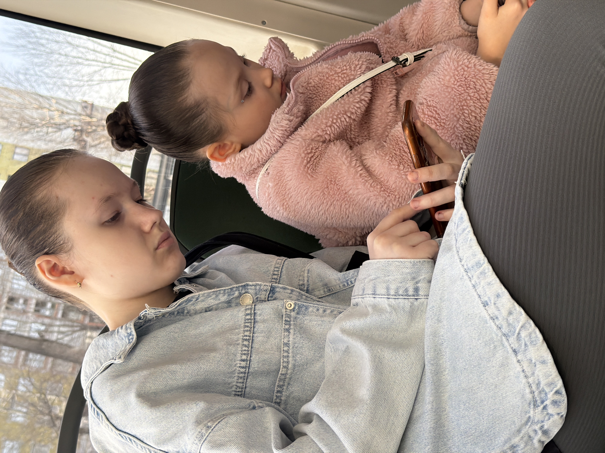 Two girls are sitting on a seat inside what appears to be a bus. The location, based on the prompt, is Perm, Russia. One girl, wearing a light blue denim jacket, appears to be older and is looking to the side with her eyes closed or lowered. She is holding a brown phone in her hands. The younger girl, wearing a pink fuzzy jacket, is lying down with her head near the older girl. The lighting is soft, and the background is visible through the bus window, showing buildings and trees, indicating an urban area.