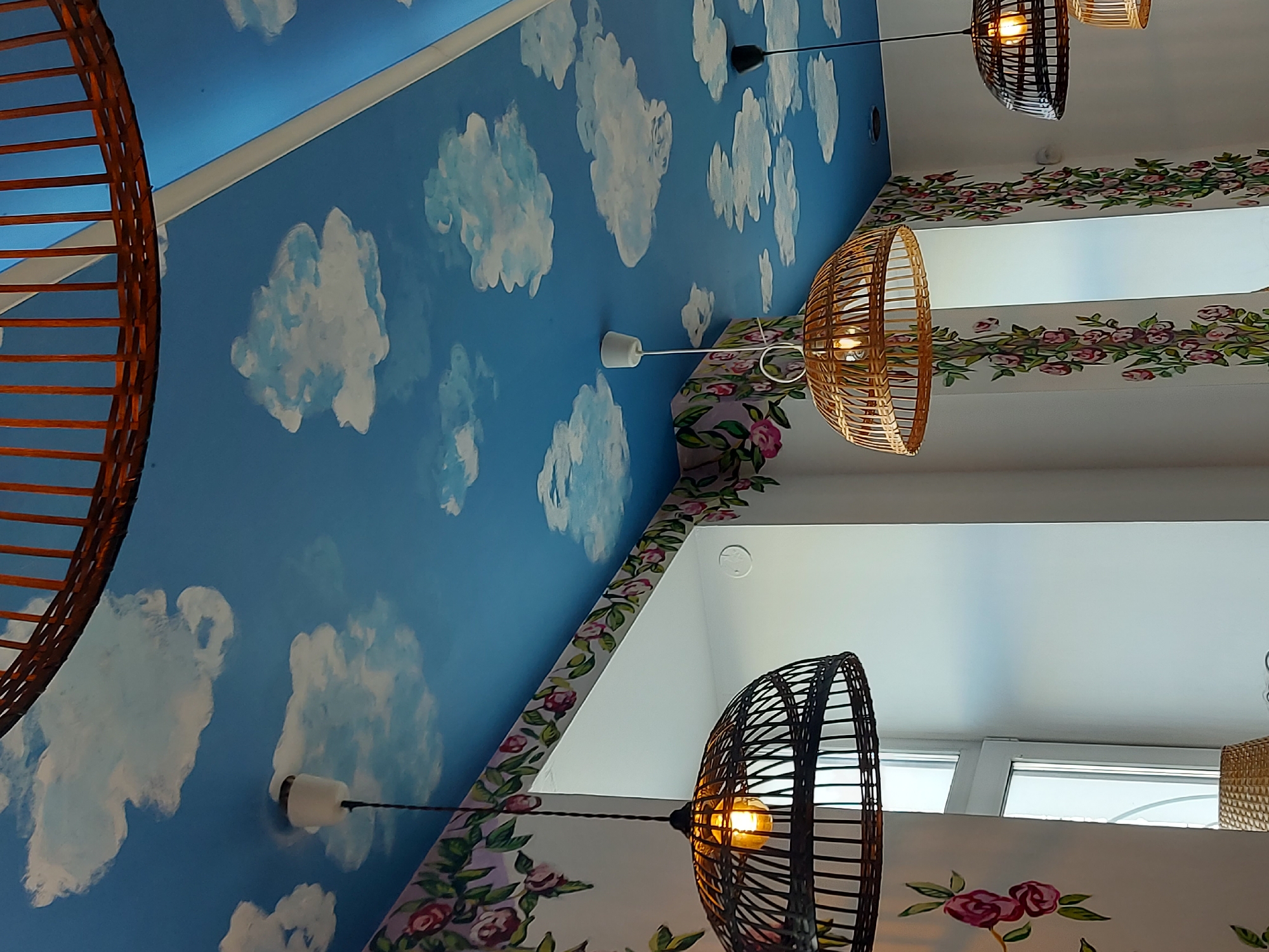 The image captures an interior view, most likely of a restaurant or cafe, in Saint Petersburg, Russia. The ceiling is divided into two distinct areas: one painted with a bright blue sky and white clouds, reminiscent of a mural, and the other is plain white. These two ceiling sections are separated by a floral design painted along the border where they meet.
Hanging from the ceiling are several woven pendant lights, some dark brown or black, and others light brown. Natural light comes in from the windows. There is a part of a large wooden structure visible on the left side. The overall scene evokes a whimsical and artistic ambiance.