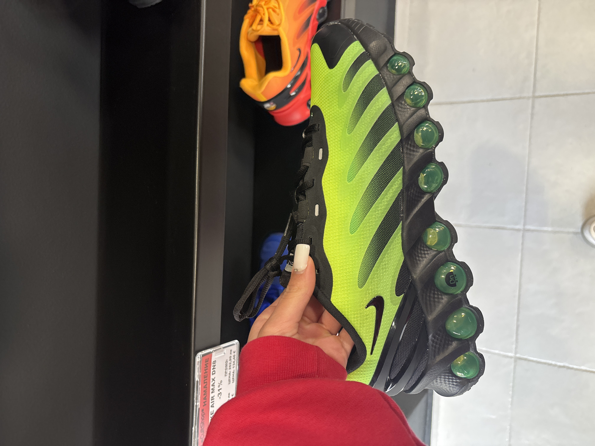 A person holds a green and black Nike Air Max DN8 sneaker with visible air bubbles, likely in a retail setting. The shoe is displayed near a shelf containing an orange and black sneaker. A price tag is visible, indicating a 31% discount for "CultShop" on the "KE AIR MAX DN8," listed in Bulgarian as "НАМАЛЕНИЕ," with a price in Euros. The location is implied as Gabrovo, Bulgaria based on the Cyrillic text on the price tag. The background consists of a dark gray display unit and a light gray tiled wall.