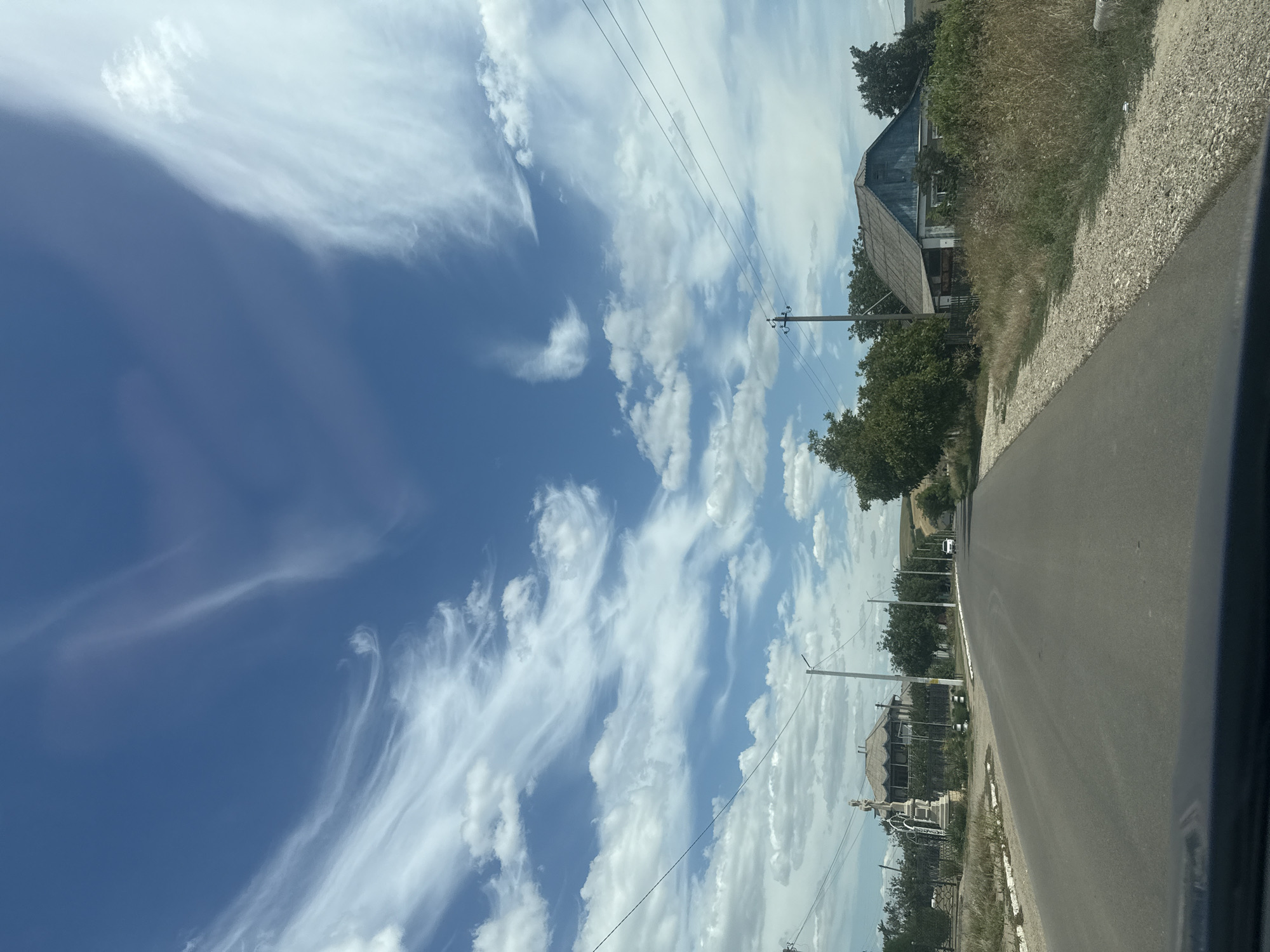 This image depicts a rural scene in Iurievca, Moldova, on a bright, partly cloudy day, likely mid-morning to early afternoon. The perspective is from a moving vehicle, looking towards the right side of the road. The foreground is dominated by a gray asphalt road that curves slightly to the right. To the right of the road, there is a gravel shoulder and a grassy verge with dry, tall grass.

Further down the road, several houses and trees are visible. One prominent structure is an older, two-story house with a weathered, multi-pitched roof, partially obscured by overgrown vegetation. Further along, other buildings, some appearing residential, are interspersed with green trees. Utility poles with visible wires run along the side of the road, suggesting the presence of infrastructure.

The sky is a prominent feature, with a vibrant blue expanse filled with various cloud formations. Wispy, cirrus clouds stretch across the upper left of the frame, creating a dramatic effect against the deeper blue. Puffy cumulus clouds are scattered throughout, indicating fair weather conditions.

No people are explicitly visible in the image. The scene evokes a sense of quiet, everyday life in a rural setting. The lighting suggests it is daytime, with sunlight illuminating the landscape. The overall impression is one of a slightly weathered but functional rural community under a dynamic sky.