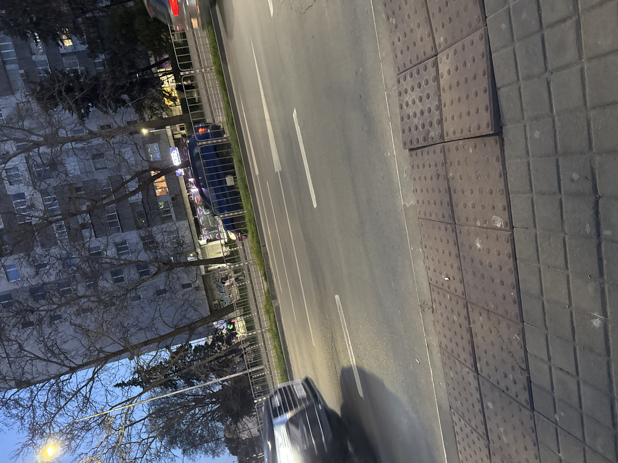 The image depicts a street scene in Varna, Bulgaria, with a gray road extending from the top to the bottom of the frame. A white solid line divides the road into lanes. A sidewalk with textured paving stones is visible on the right side. A building, likely an apartment block, stands on the left side of the street, partially obscured by bare trees. A metal fence runs along the left edge of the road. Cars are on the road, one is closest to the bottom of the image. The sky is visible as dusk or dawn, providing a soft, low-light atmosphere. There are no pedestrians visible in the frame.