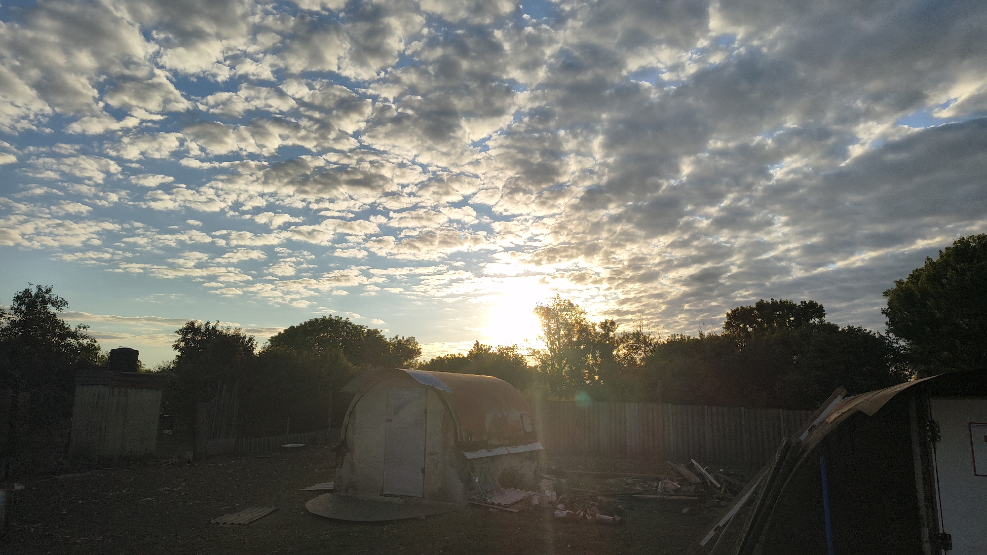 The image depicts an outdoor scene at sunrise or sunset, with the sun low on the horizon and casting a warm glow through scattered clouds. The foreground shows a dirt or gravel ground with some debris, including what appear to be pieces of wood and roofing material. There are a couple of small structures; one is a light-colored shed with a dark roof, and another is a more prominent, rounded structure with a reddish-brown roof and a white door. A corrugated metal fence runs horizontally across the midground, separating the area with the structures from the background. The background is dominated by a dense line of trees, silhouetted against the bright sky. The sky itself is a mix of blue and white, with the sun positioned slightly left of center, peeking through the clouds and creating lens flare effects. There are no people visible in the image.