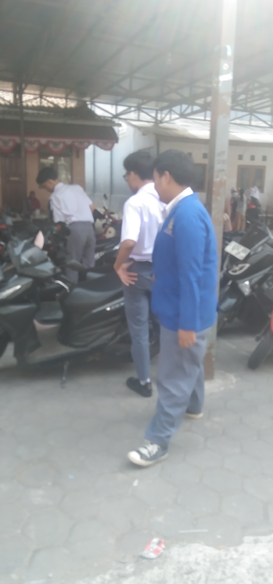 In the city of Rahayu, Indonesia, outside a building with a corrugated metal roof supported by metal beams, three individuals are present. The foreground features two young males, likely students based on their uniforms. One is wearing a white short-sleeved shirt and grey pants, with his back to the camera, leaning over a dark-colored motorcycle. The second individual, to his right and slightly forward, wears a blue jacket over a white shirt and grey pants, facing towards the right side of the frame. He has his right hand resting on his hip. A third young male, wearing a white shirt and grey pants, stands behind the first, also with his back mostly to the camera. Both of the foreground individuals are wearing dark-colored athletic shoes. Several dark-colored motorcycles are parked in the paved area, which has a hexagonal tile pattern. The environment is bright, with sunlight casting a glare on the scene, particularly from the upper right. The background shows parts of buildings, including a red and white striped awning over a window.
