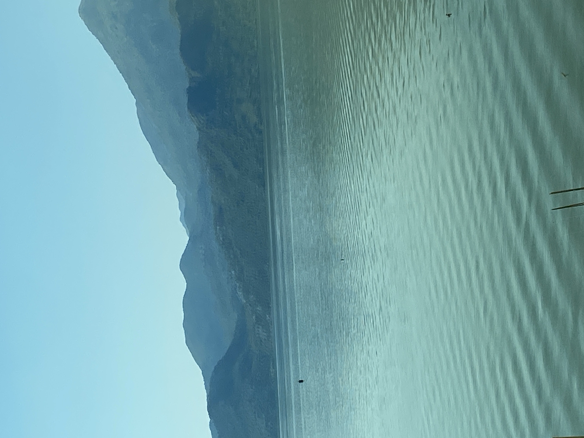 The image depicts a serene natural landscape featuring a wide body of water, likely Lake Skadar, with rolling hills and mountains in the background. The sky is a clear, pale blue, suggesting a calm and bright day. The water has a tranquil surface with gentle ripples, reflecting the muted tones of the surrounding hills. A dark, rectangular object, possibly a buoy or a small marker, is visible in the water towards the left side of the frame. The overall impression is one of peaceful solitude and natural beauty.
