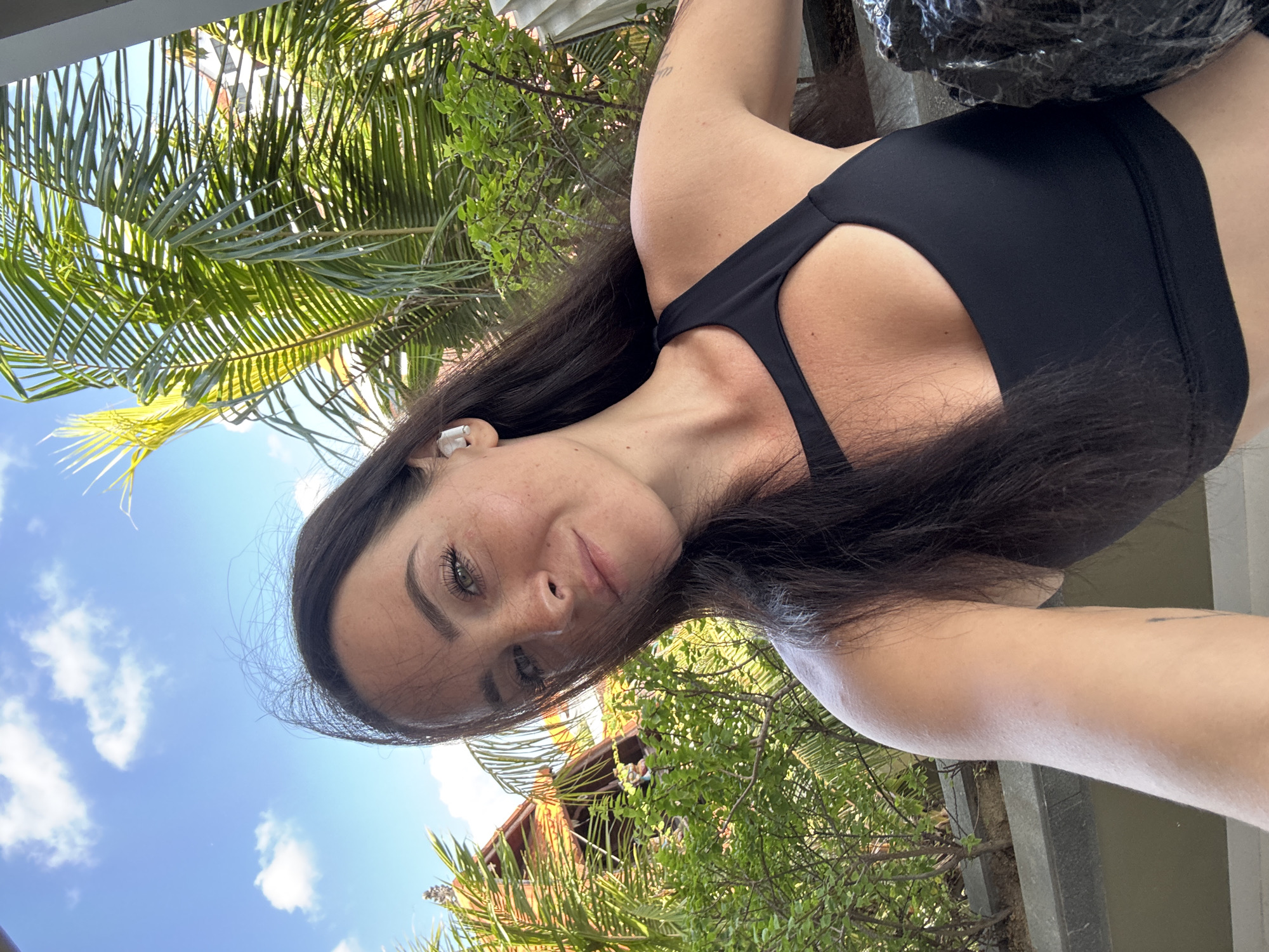 A woman with long, dark hair and fair skin takes a selfie outdoors. She is wearing a black sports bra and white wireless earbuds. Her hair is partially obscuring her face, and she is looking at the camera with a slight smile. Behind her, there are palm trees, lush green foliage, and a building with an orange tiled roof, suggesting a tropical or subtropical location. The sky is blue with scattered white clouds. The overall impression is one of a casual, relaxed moment in a warm climate, possibly at a resort or in a residential area in North Kuta, Indonesia.