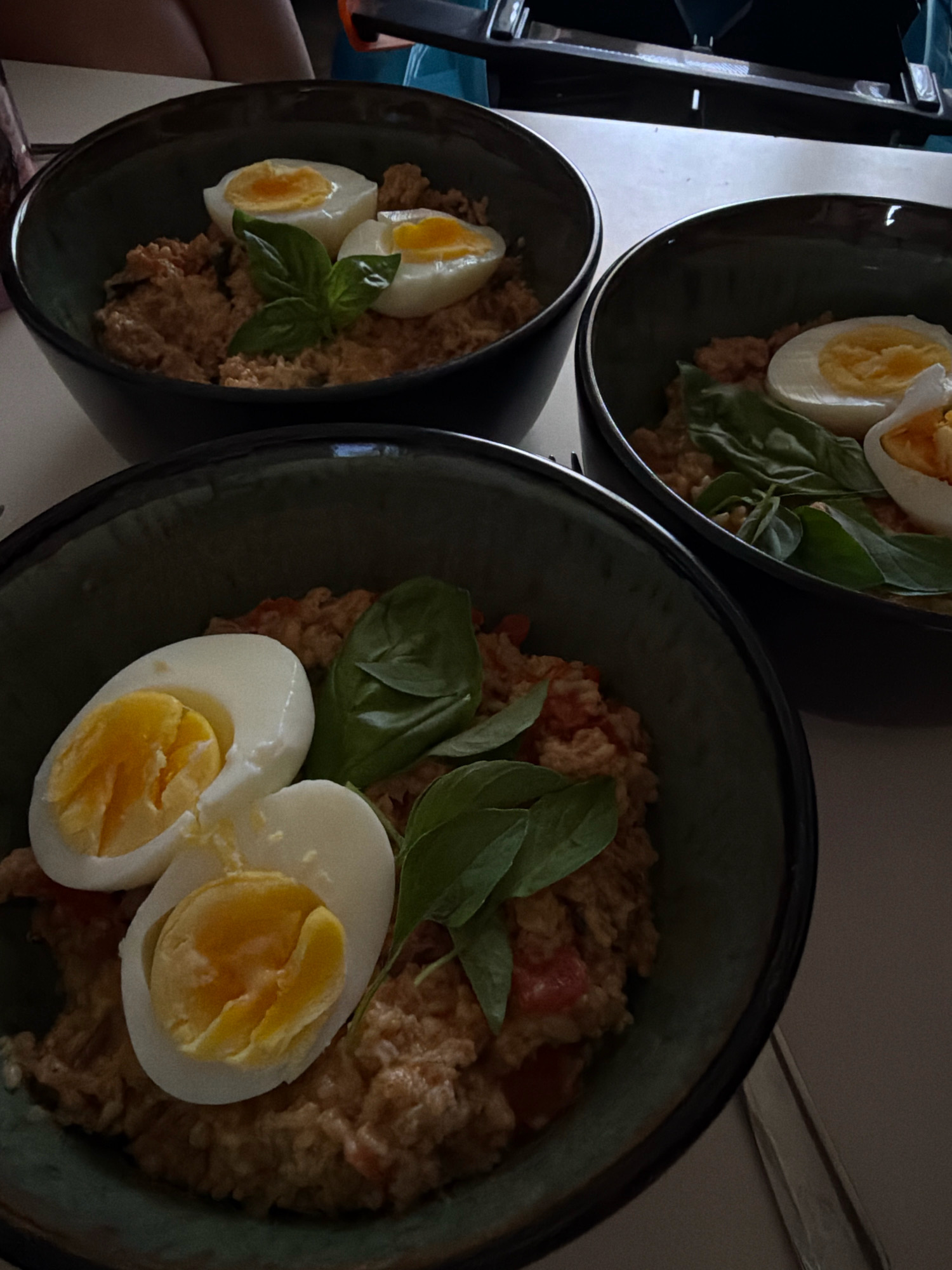 The image displays three bowls of food, each containing a portion of what appears to be rice or a grain-based dish mixed with shredded meat or fish, and visible pieces of tomato. Each bowl is garnished with two halves of a hard-boiled egg and a sprig of green leaves, likely basil. The bowls are dark green on the outside and have a rough texture. One bowl is in the foreground, and two are positioned behind it. The lighting suggests an indoor setting, possibly a restaurant or dining area. The background is blurred, showing a tabletop and a person's legs in dark clothing. There are no discernible landmarks or elements that would definitively place the image in Sharm El Sheikh, Egypt.