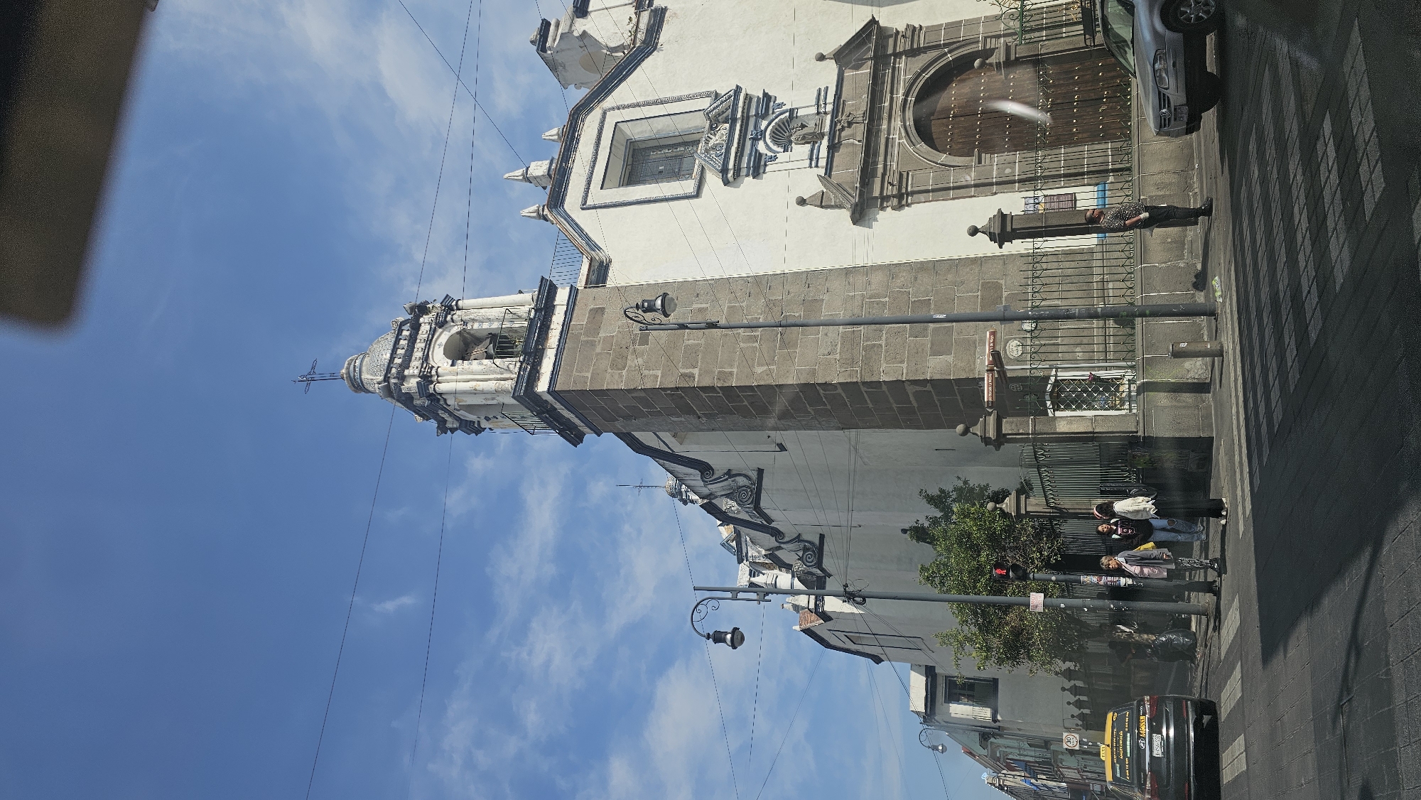 The image captures a vibrant street scene on a sunny day in what appears to be a historic town, featuring a prominent colonial-era church.
The main subject is a large, ornate church building dominating the left side of the frame. Its lower section is constructed from grey stone blocks, while the upper facade and bell tower are white with intricate carvings and blue and white decorative tiles. The bell tower features a domed top crowned with a cross. A large arched wooden door serves as the main entrance, and a rectangular window above it is adorned with detailed stonework. A fence separates the church property from the sidewalk.
On the paved street and sidewalk, daily activity is visible. A silver car is parked near the church entrance. Several individuals are present: one person in a patterned shirt stands near the fence, while a group of three people, possibly young women, are further down the street, near a traffic light and a marked crosswalk. A dark-colored taxi with a yellow roof light is visible in the bottom right, with "TAXI" and a license plate (partially visible as "B38-662") on its side.
The setting includes streetlights with decorative scrolls, green foliage from trees or bushes, and utility wires crisscrossing the clear blue sky, which has scattered white clouds, indicating daytime. A small brown sign on a pole near the church is visible but illegible. The architectural style and street elements are consistent with the location context of Puebla, Mexico. The scene depicts a typical bustling street environment with no specific event unfolding.
