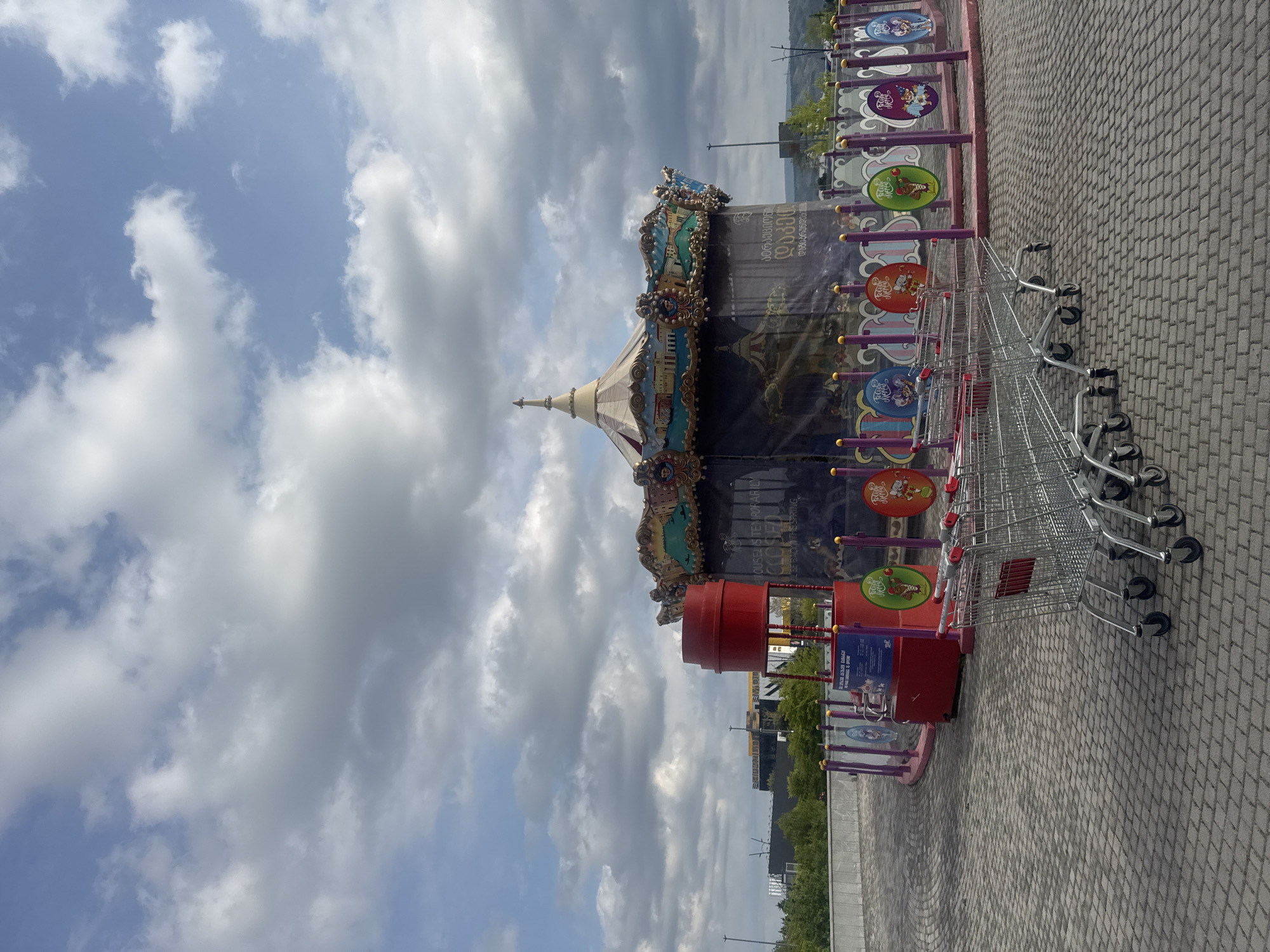 The image, oriented sideways, displays an outdoor scene featuring a carousel and a line of shopping carts under a partly cloudy sky. The carousel, located on the left side of the frame when viewed upright, is large and ornate, featuring a striped, tent-like roof and decorative panels with painted scenes and gold accents. A blue and purple banner wraps around the carousel's base, displaying Georgian text that translates to "Carousel Temporarily Closed Until Spring," indicating it is not operational.
Adjacent to the carousel, a row of silver metal shopping carts with red handles is parked on a grey paved surface made of interlocking bricks. A purple barrier or fence, adorned with several colorful circular signs depicting various cartoon characters, runs alongside the carts and the carousel area. One red sign attached to this barrier also contains text in both English and Georgian, stating "CAROUSEL RULES AND REGULATIONS."
The sky above is bright, with significant cloud cover interspersed with patches of blue. In the background, hills or mountains, trees, and distant structures are visible. No people are present in the scene. The combination of a carousel and shopping carts, along with the visible Georgian text, suggests a commercial or recreational area, likely a shopping center or park, in Tbilisi, Georgia. The time of day appears to be daytime, with clear visibility.