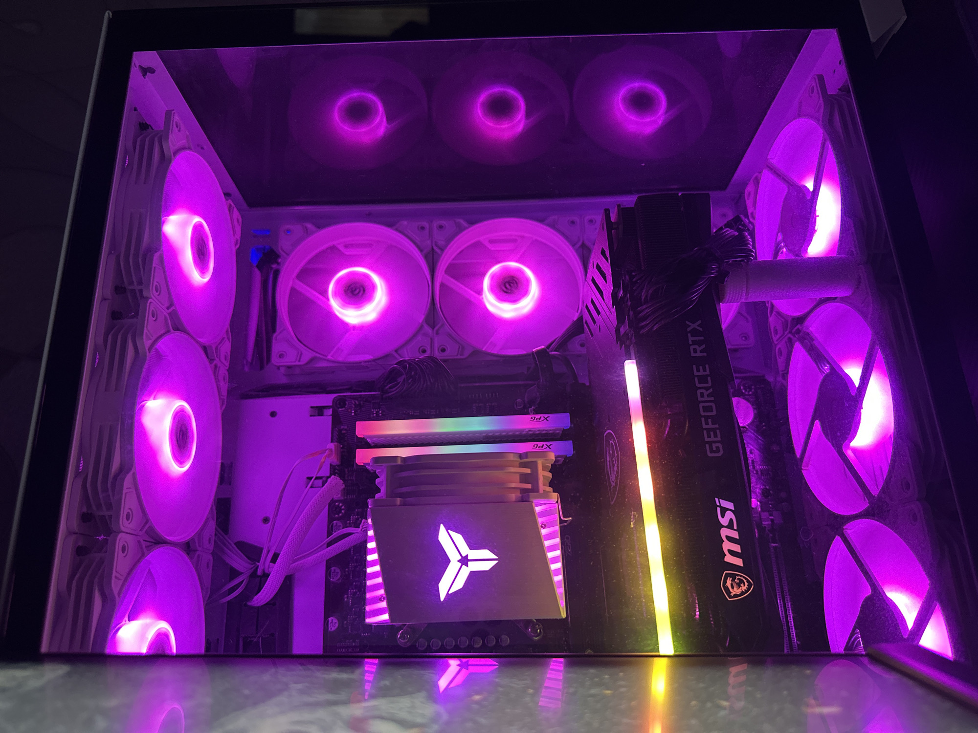 The image shows the interior of a computer case, illuminated by purple and pink LED lights. The case is likely a gaming PC, given the visible components and aesthetic. Multiple fans, some on the side and some on the top, are lit up with vibrant purple light, creating a futuristic and energetic atmosphere. A large graphics card, an "MSI GEFORCE RTX," is prominently featured, also glowing with RGB lighting. A CPU cooler with a distinctive "Y" shaped logo is visible in the center, with its own illuminated accents. Two RAM sticks with "XPG" branding are installed on the motherboard. The overall scene suggests a powerful and visually striking computer setup, likely in a dark environment to emphasize the lighting effects. The reflection of the components on the surface below hints at the case possibly having a glass bottom or being placed on a reflective surface. No people are visible, and the focus is entirely on the internal components and their lighting.