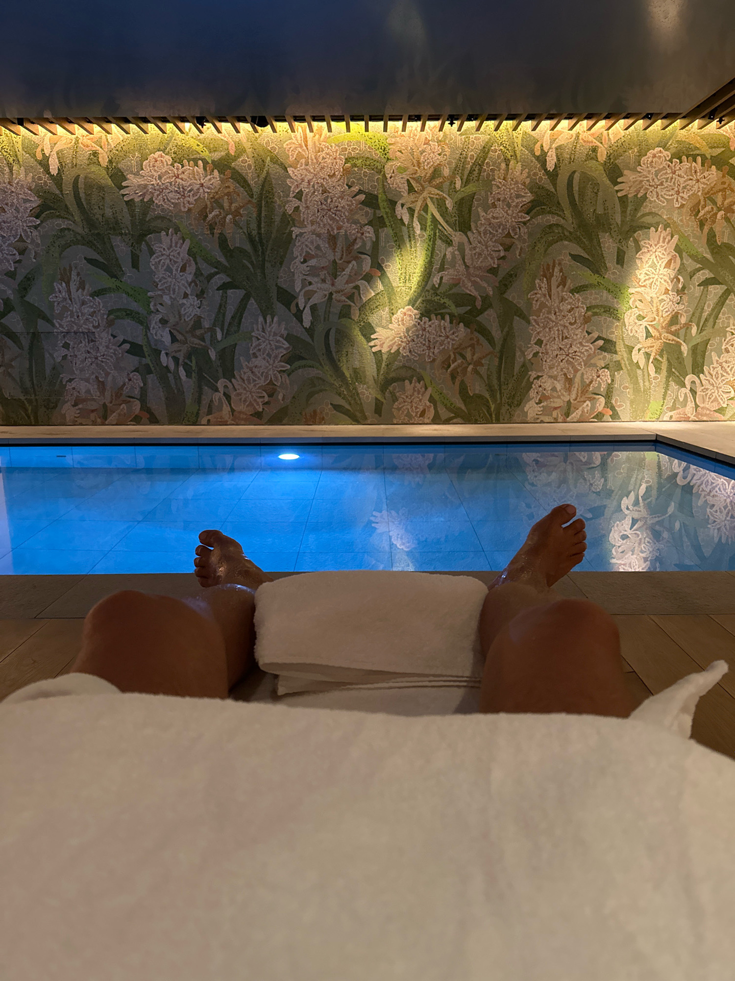 A first-person perspective shot shows a person's legs and feet resting on a white towel, with a small indoor swimming pool and a floral tiled wall in the background. The person's legs are covered with a towel, but their feet are visible, appearing wet. The pool water is a bright blue, and the wall behind it features a mosaic of light-colored flowers against a green background, illuminated by warm lighting. The scene appears to be indoors, possibly in a spa or hotel. There is no indication of the location being in London, United Kingdom.