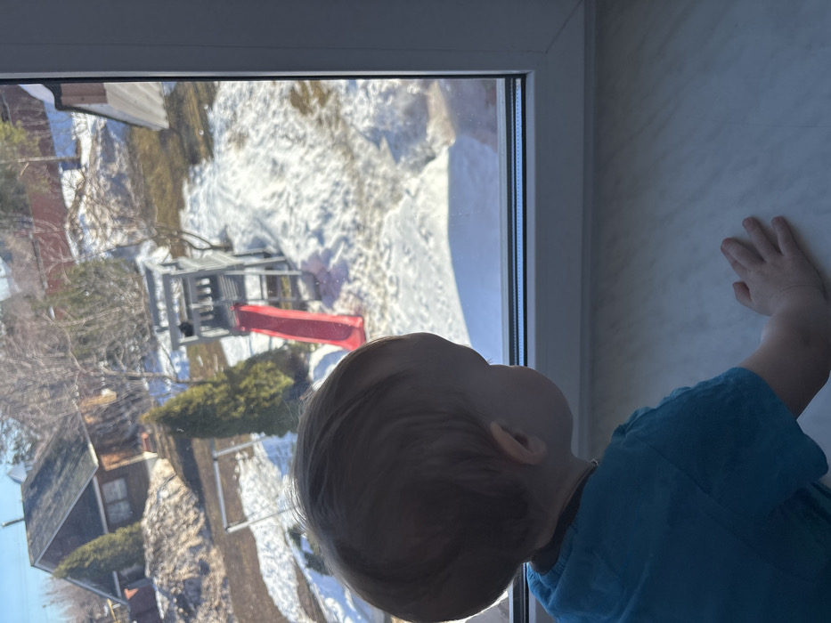 A young child, with light brown hair and wearing a short-sleeved blue top, is positioned with their back to the viewer, looking out of a white-framed window. The child's left hand rests flat on a light-colored, possibly marbled, windowsill. The scene outside the window, located in Shul'gino, Russia, depicts a residential backyard environment under bright daylight, indicative of late winter or early spring. The ground is partially covered in melting snow, interspersed with visible patches of brown grass and bare earth. A prominent wooden playset, featuring a red slide and a ladder, is situated in the middle ground. Various trees, including bare deciduous trees and green coniferous trees, are present. In the upper left background, a small, dark-roofed, brown house is visible behind a large pile of earth or debris. Distant fencing marks property boundaries.