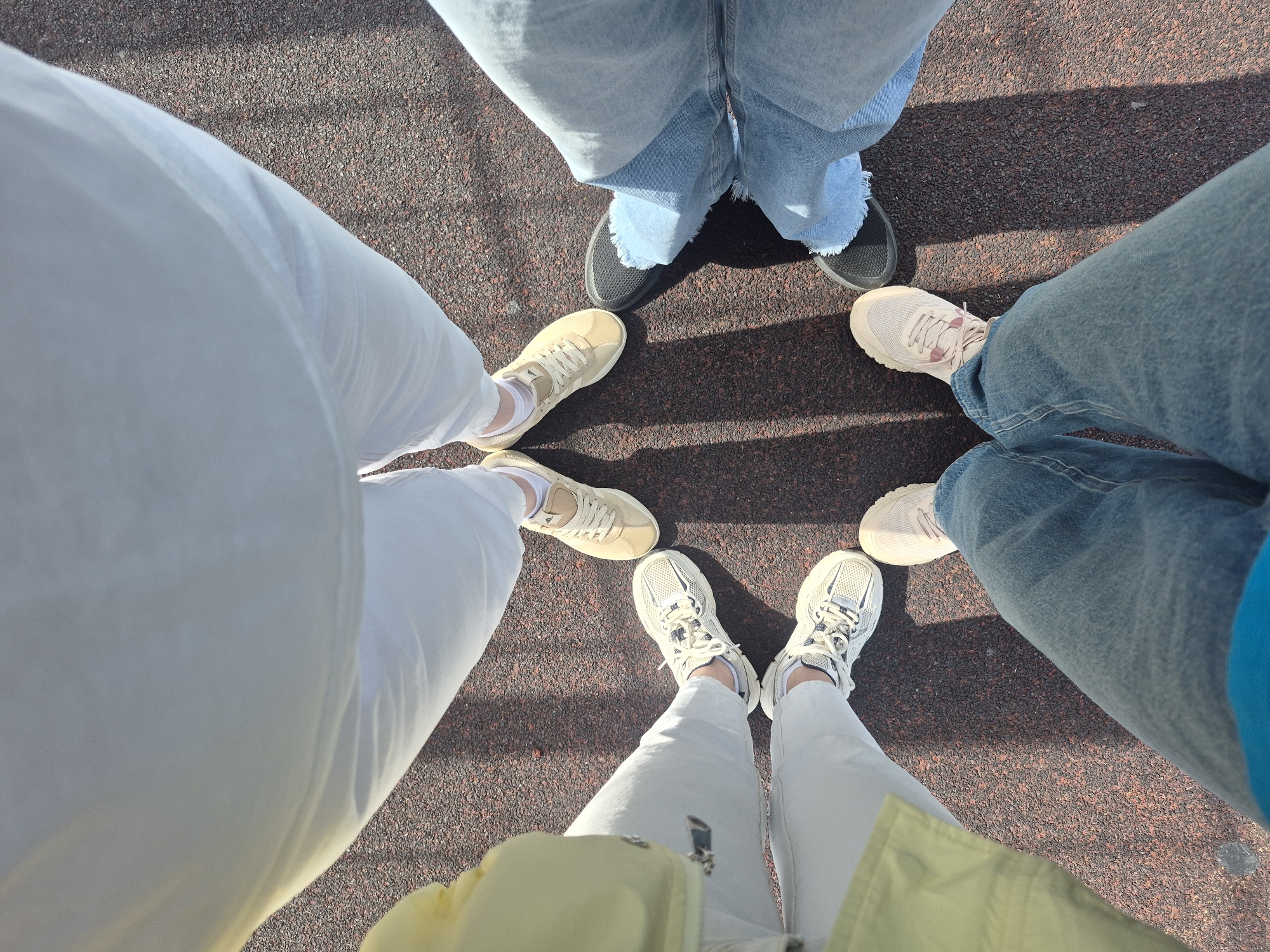 A high-angle shot shows five pairs of legs and shoes standing on a dark red surface. One pair wears light-colored jeans and tan sneakers. Another pair wears blue denim jeans and black shoes. A third pair wears light blue jeans and pink sneakers. A fourth pair wears white pants and white sneakers. The last pair wears white pants and cream-colored sneakers. The location is Unknown, Unknown.