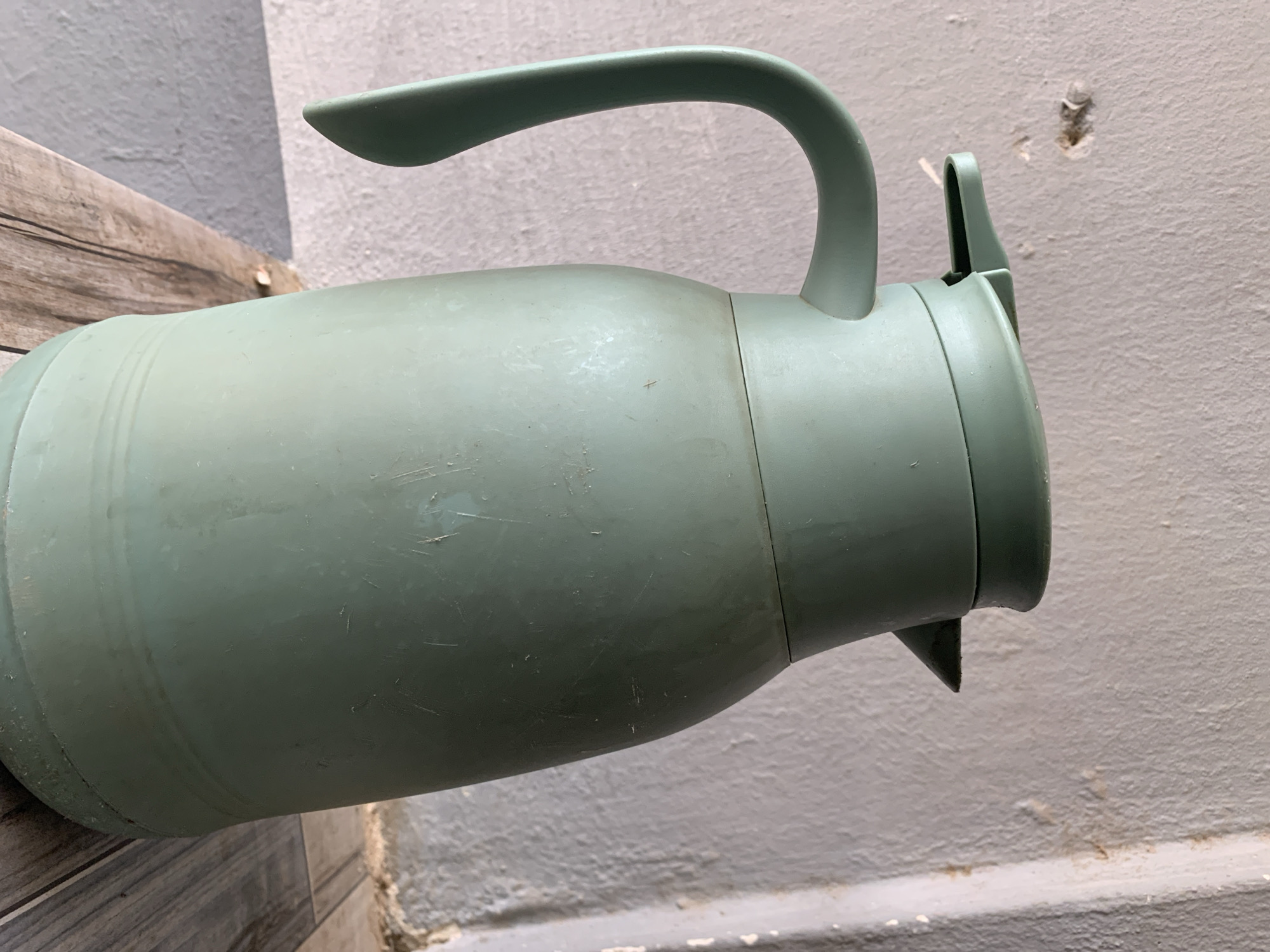 A light sage-green plastic thermos flask is centered in the frame, slightly angled to the right. The flask features an integrated handle on its left and a covered pouring spout on its right. Its surface displays multiple linear scratches, scuff marks, and dust accumulation, most notably around its lower section and base. The background consists of a textured, off-white wall, which presents a circular plaster defect approximately 3 cm in diameter on the upper left, revealing a darker substrate. To the right, a vertical architectural edge is visible, accompanied by faint, irregular brown-orange discolorations on the wall surface. The thermos rests on a horizontal surface composed of greyish-brown ceramic tiles patterned to resemble wood grain. A darker, solid grey, textured surface is partially visible below the wall and to the left of the tiled surface. The scene is illuminated by diffuse light, and no individuals are present. This scene is located in New Stadium, Nigeria.