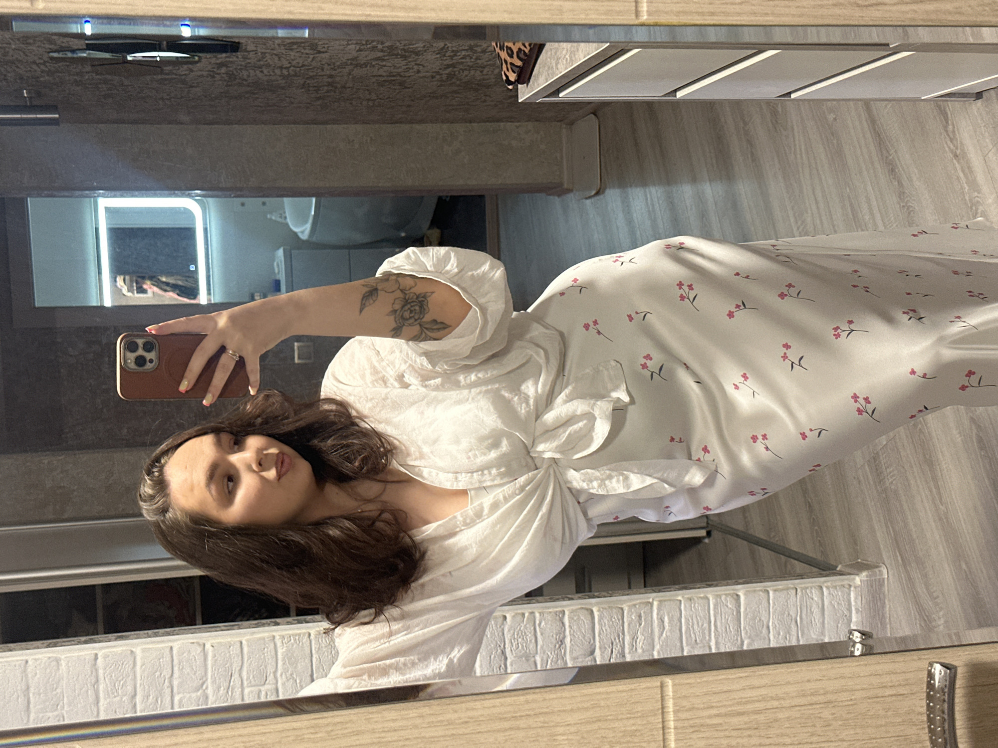 A young woman with brown hair is taking a selfie in a bathroom mirror. She's holding a brown smartphone in her left hand. She is wearing a white wrap dress with a small pink floral print. A tattoo is visible on her left forearm. The bathroom features a light-up mirror above a vanity. The mirror reflects a toilet. The location is Chaikovsky, Russia.