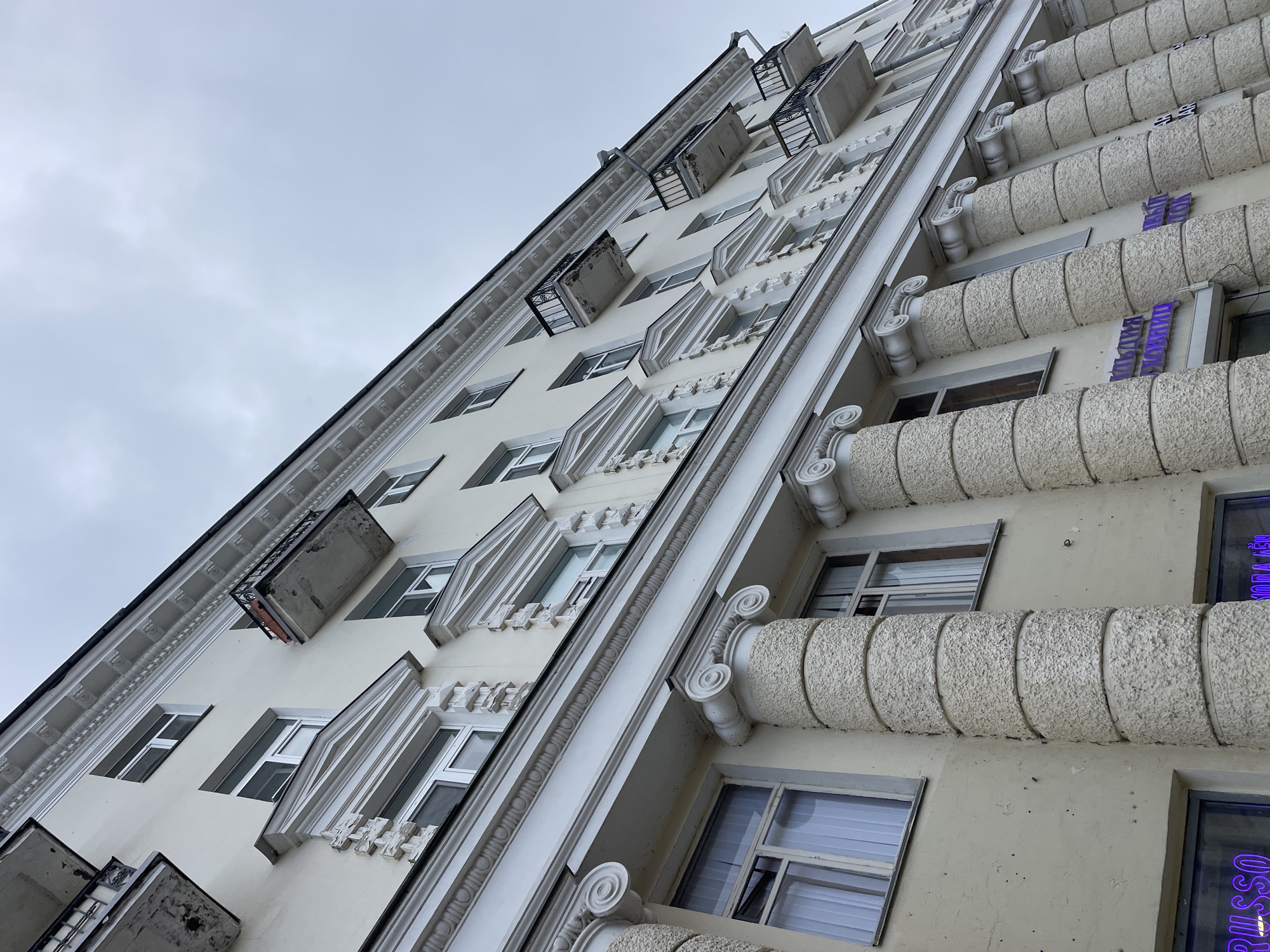The image presents a low-angle, upward-looking view of a multi-story building facade in Kazan, Russia, against a uniformly overcast, light gray sky. No people are visible.
The building exhibits a blend of classical and Art Deco architectural styles, predominantly in a light cream or off-white color with crisp white decorative elements. On the right side of the frame, the facade features several vertical rows of rectangular windows, interspersed with heavy, horizontal bands of rough-textured concrete. These bands are supported by ornate, scrolled capitals, creating a robust, column-like aesthetic. Two purple neon signs are prominently displayed on this section. The upper sign clearly spells "ВИШНЕВЫЙ" (Vishnevyy, meaning "Cherry"), while the lower one partially reveals "РУССО" (Russo).
Towards the left, the building's design shifts to a more refined, yet still decorative, style. This section features numerous windows set within recessed, white-trimmed frames. Small, projecting metal balconies with intricate railings punctuate the facade; some appear shallow or purely ornamental. A strong vertical white molding, adorned with a repeating scroll motif, runs along the building's edge, enhancing its classical feel. A simple downspout is also visible near the top left. The consistent gray sky suggests an overcast day, possibly cool and muted. The architecture points to a historic or well-preserved structure, likely an apartment building or commercial establishment.