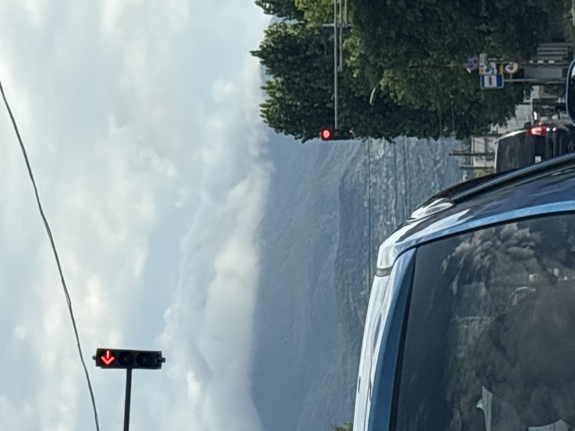 The image shows a view from inside a car, looking out towards a mountain range partially obscured by clouds. In the foreground, the blue roof and side of a car are visible. A traffic light with a red left arrow is illuminated, mounted on a pole extending from the left side of the frame. Further in the distance, another traffic light with a red circular signal is visible amidst dense green trees. Several other cars and a street sign are also present, suggesting an urban environment. The scene captures a moment in Tbilisi, Georgia, likely on a day with variable weather conditions affecting visibility of the mountains.
