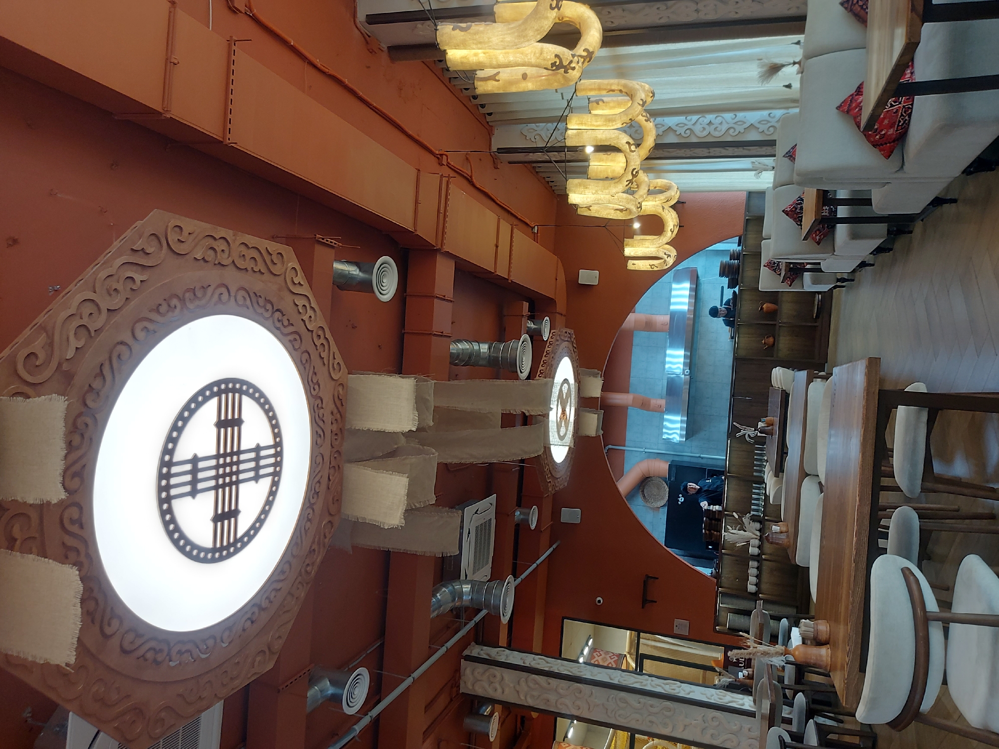 The image depicts the interior of a restaurant with a warm and rustic ambiance. The ceiling is painted a vibrant orange and features decorative wooden carvings and suspended fabric panels. A large, circular light fixture with intricate geometric patterns is a prominent ceiling element. Several industrial-style air vents and metal pipes are visible, adding a contrasting utilitarian touch.
The restaurant is furnished with wooden tables and chairs, as well as comfortable booth seating with upholstered cushions adorned with decorative patterns. Two chandeliers with a unique, organic design hang from the ceiling, emitting a soft, warm light. In the background, a kitchen area is partially visible, with a chef in uniform working behind a counter. The flooring appears to be light-colored wood laid in a chevron pattern. The overall impression is of a thoughtfully designed space that blends traditional elements with modern industrial aesthetics.