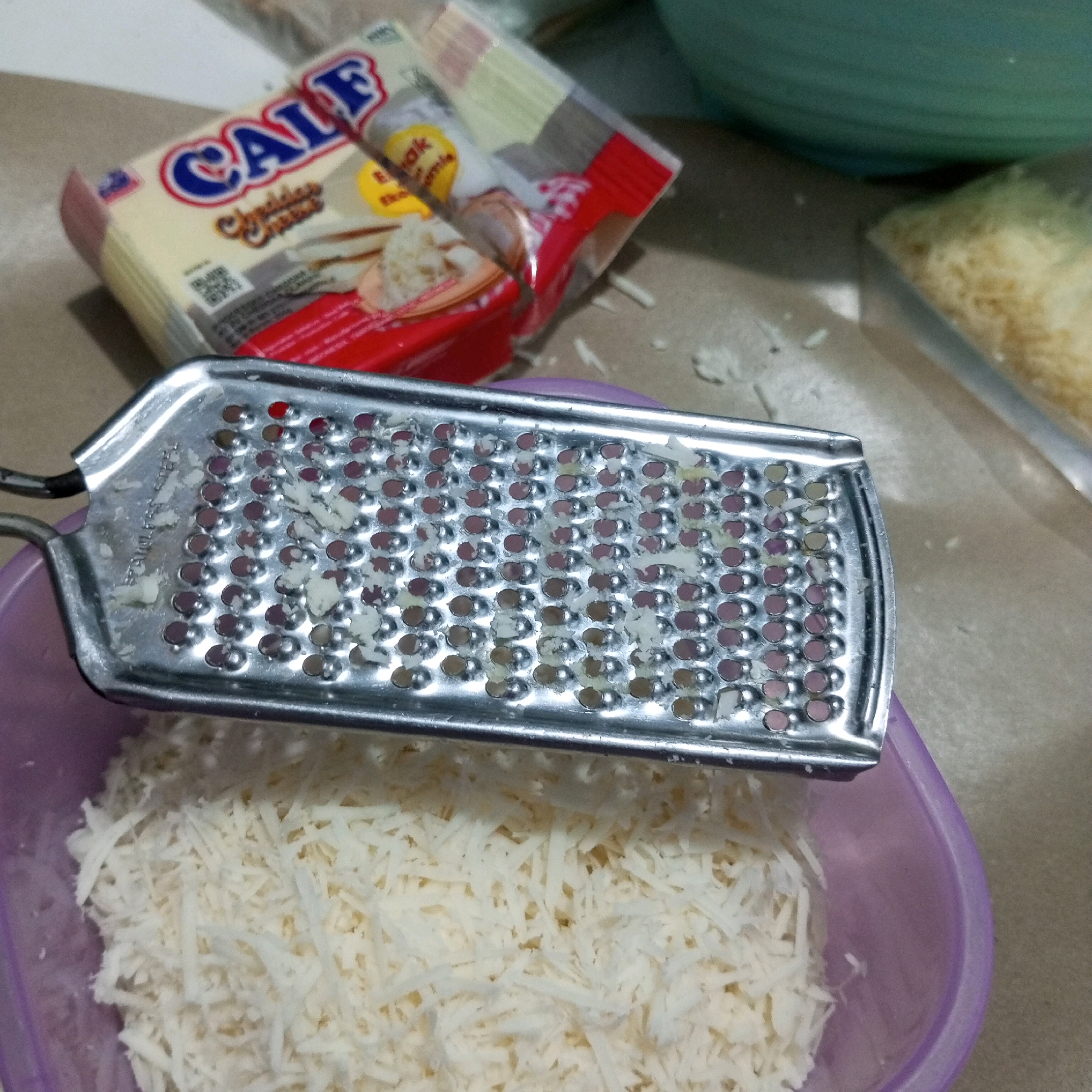 In a kitchen setting, likely in North Jakarta, Indonesia, a block of "Calf" brand cheddar cheese is partially visible, suggesting it is about to be grated. A metal grater, stained with remnants of grated cheese, rests on the edge of a purple plastic bowl. The bowl is filled with a generous amount of finely grated cheese. The overall scene depicts the preparation of cheese for cooking or a meal.