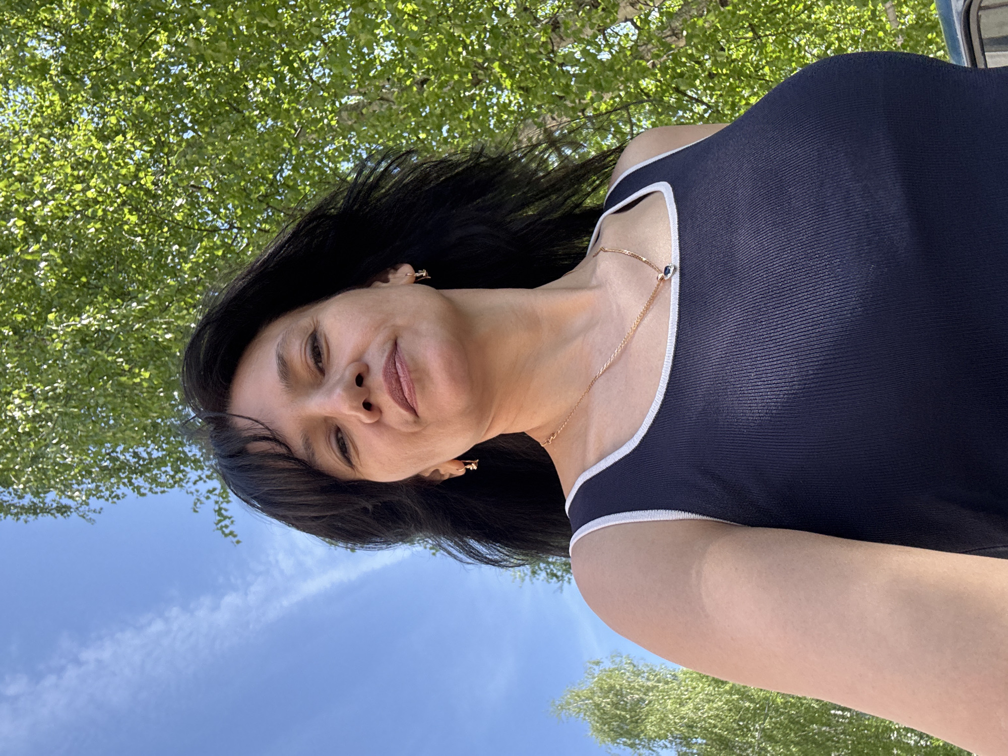 A low-angle shot looking up at a woman. She has dark hair, light skin, and is wearing a dark blue tank top with white trim. She also has a thin gold necklace with a small pendant and gold earrings. The background shows a blue sky with wispy clouds and green foliage from trees. The location appears to be outdoors, possibly a park or wooded area. The city of Chaikovsky is not identifiable from the image.