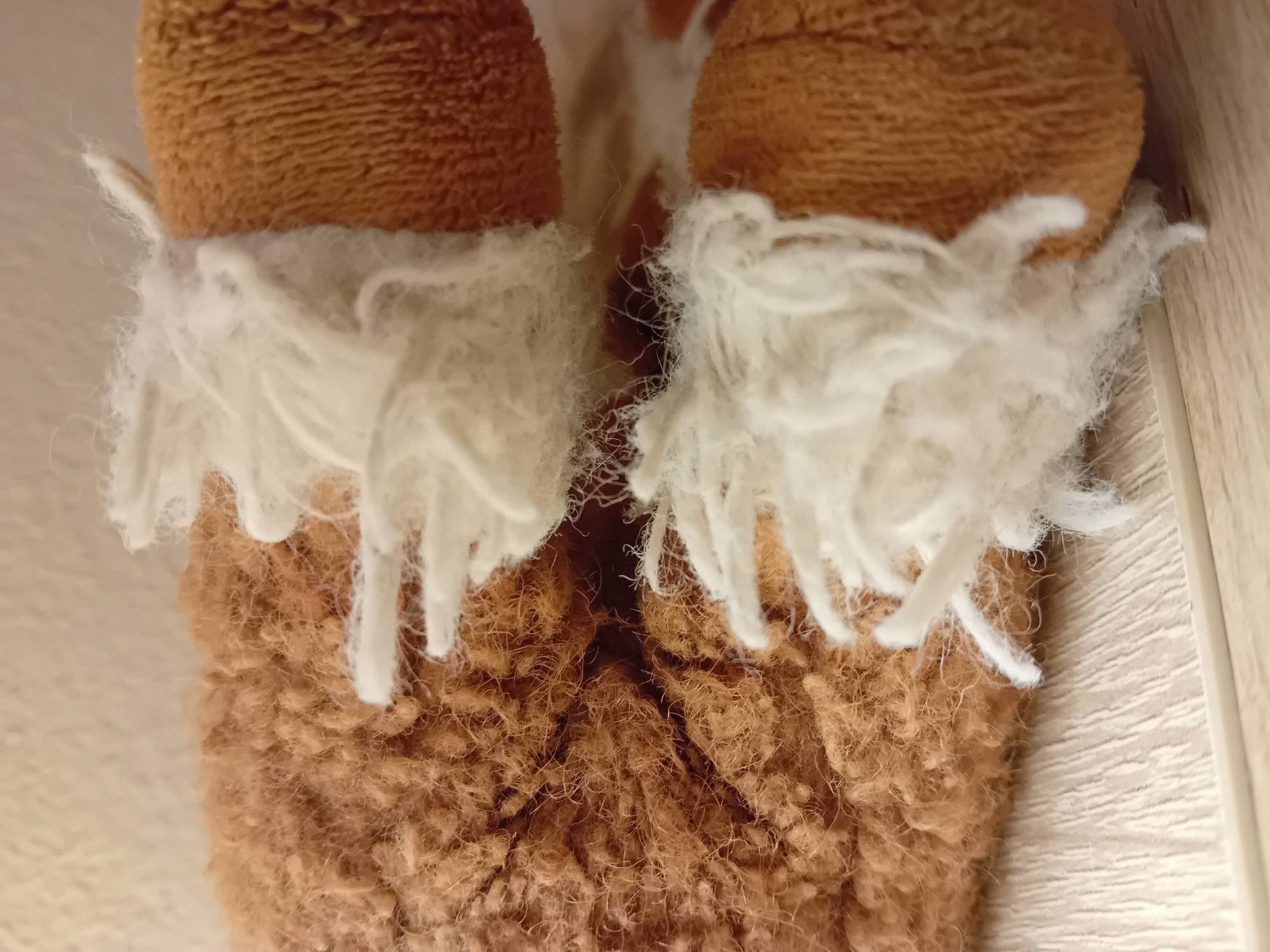 The image shows a close-up of a plush toy, likely a stuffed animal. The toy has sections of brown, fuzzy material and sections of white, fluffy material that resembles hair or fur. The brown material appears to be a coarser, matted texture, while the white material is wispy and loose. The toy is resting on a light-colored, textured surface, possibly wood grain, with a thin white border at the bottom. The lighting is soft and casts subtle shadows. There are no people or identifiable landmarks in the image, and no specific location in Zuberec, Slovakia, can be determined from the visual information provided.