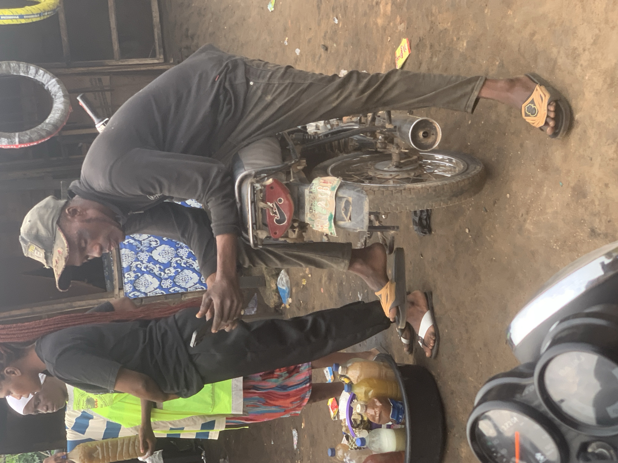 In the city of Shagamu, Nigeria, a man wearing a cap and a black long-sleeved shirt and pants is perched on a motorcycle, leaning his elbows on his knees. His right hand is resting on his left hand. He is looking down and to his left. To his left, another person wearing a black t-shirt and black pants is standing with their hands clasped in front of them, also looking down. Between them, a person in a green vest and a brightly colored skirt holds a clear plastic bottle filled with a brownish liquid. In the foreground, a dark plastic basin holds several bottles of liquid, some clear and some brownish. Behind the people, a shop appears to be selling tires, with two visible hanging from the ceiling. The ground is dirt, and there is debris scattered around. The overall scene appears to be a marketplace or roadside stall.