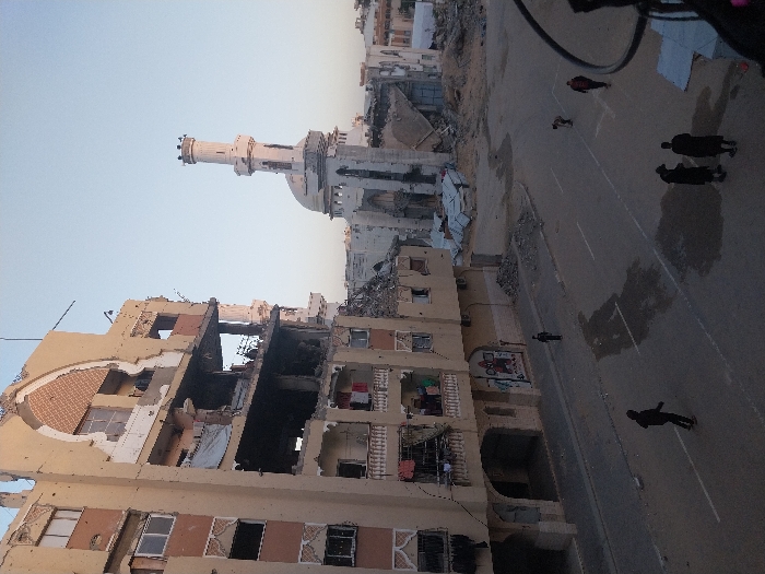 Buildings stand destroyed, likely from conflict, exposing interiors and broken facades. A mosque's minaret rises above rubble. A wide road runs alongside the buildings with several pedestrians. The scene conveys devastation and urban decay, with figures moving amidst the destruction. Dust or smoke hangs in the air, and the sky is pale.