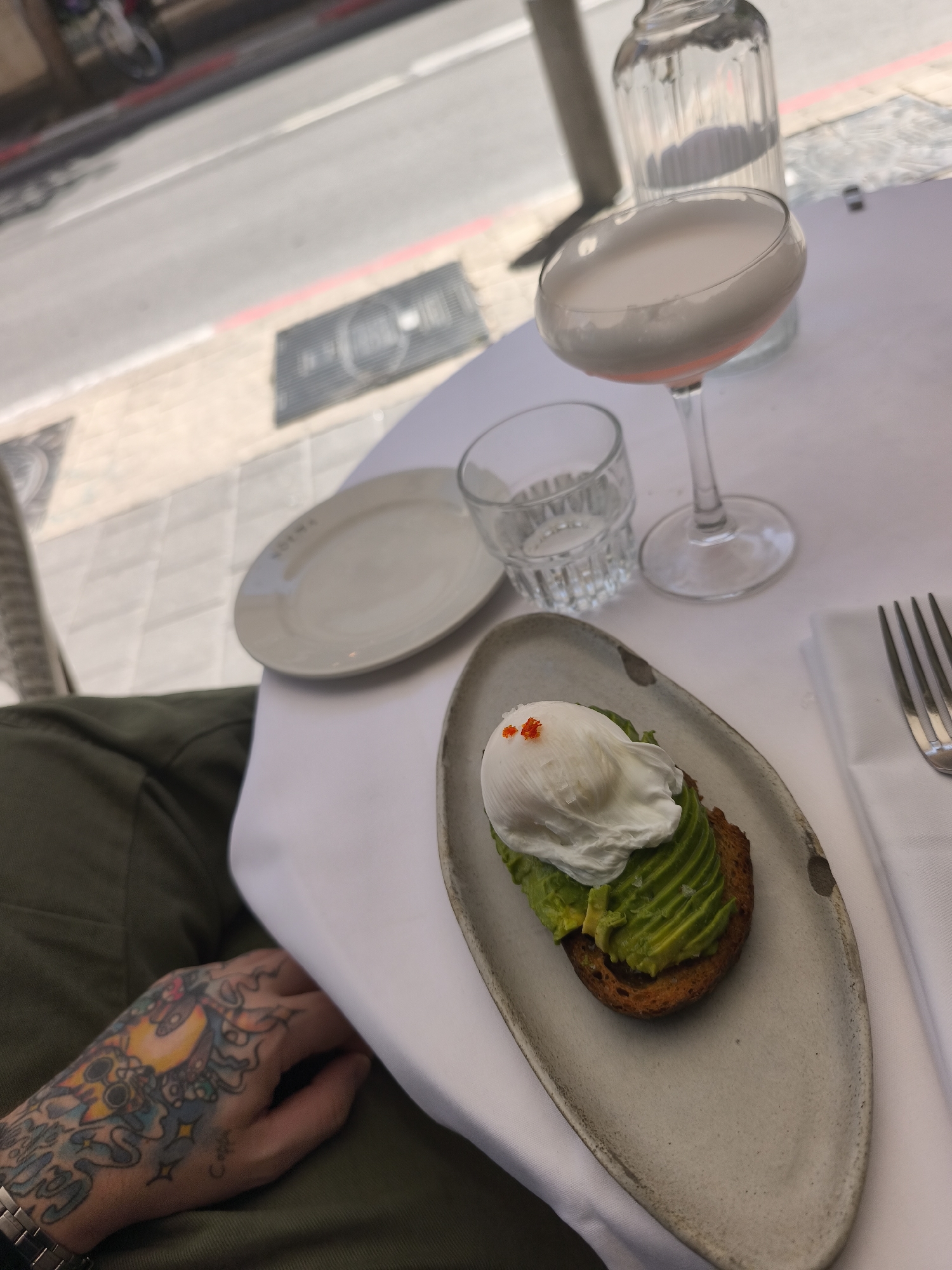 The image depicts a high-angle, slightly oblique view of a table setting at an outdoor restaurant. On a white tablecloth, an elongated, irregular light gray ceramic plate holds avocado toast topped with sliced avocado, a poached egg, and red roe. To its right, a stemmed coupe glass contains a pale pink, foamy cocktail. Behind the cocktail is a clear, fluted glass bottle filled with water. To the left of the cocktail, a clear, textured tumbler holds water. Further left, a small, white ceramic plate displays the word "COLIMA" on its rim. A silver four-tined fork rests on a folded white napkin to the right.
On the far left, the lower portion of a person's left arm and hand are visible, dressed in olive-green fabric. The hand and wrist are extensively covered with colorful, illustrative tattoos, and a silver-colored metal bracelet watch is worn on the wrist.
The background is a blurred view of a city street and sidewalk. The light gray pavement features a red painted line and several dark gray metal grates. One grate prominently displays the words "COLIMA" and "TEL-AVIV". A blurry bicycle or scooter is partially visible in the distant background. A dark vertical pillar is also present, casting a shadow. The presence of "COLIMA" and "TEL-AVIV" on the plate and a street grate confirms the location as Colima restaurant in Tel-Aviv, Israel.