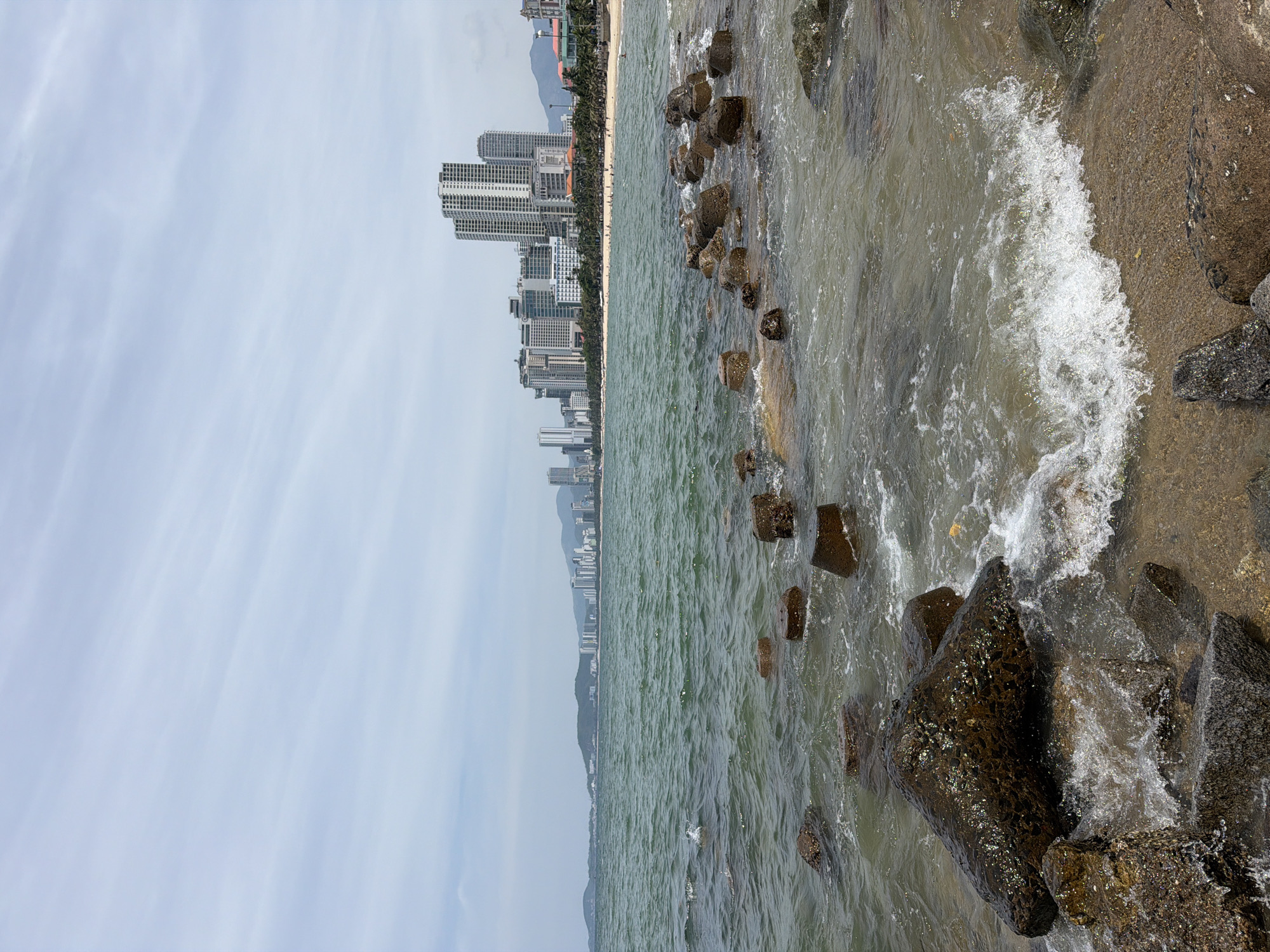 The image depicts a coastal scene in Nha Trang Ward, Vietnam, featuring a body of water with moderate waves lapping against a rocky shore in the foreground. Scattered concrete structures, likely breakwaters, are visible partially submerged in the water and along the shoreline. Beyond the water, a sandy beach stretches along the coastline, lined with palm trees and numerous tall buildings, including apartment complexes and hotels, forming a cityscape. The sky is overcast with a hazy, pale blue appearance, and distant mountains are visible on the horizon. A few individuals can be observed on the beach, and the overall impression is of a developed urban coastline on a cloudy day.