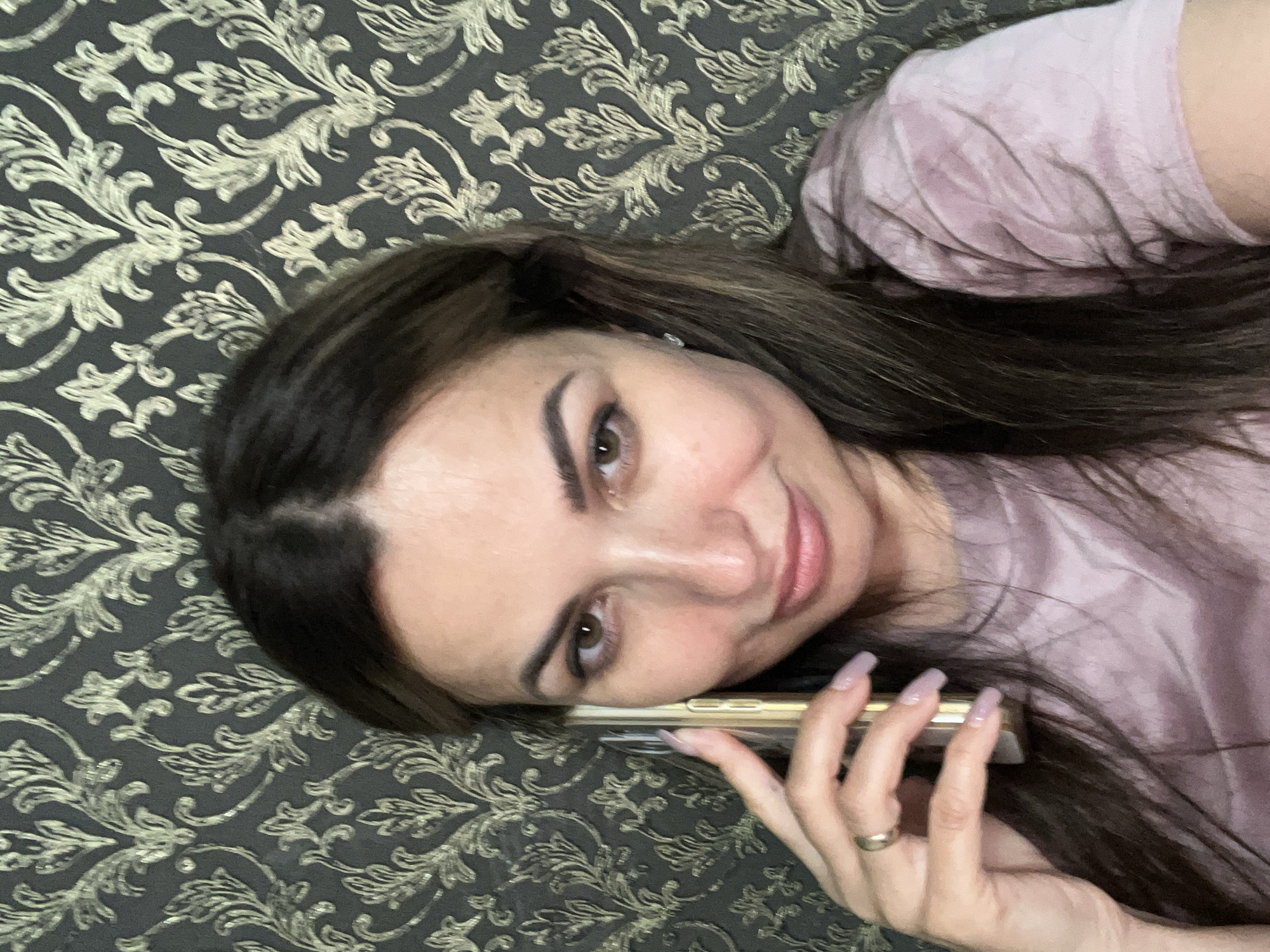 A medium shot shows a woman holding a smartphone to her left ear. She is looking towards the camera with a slight smile. The woman has long, dark brown hair and is wearing a light pink t-shirt. She has a ring on her left ring finger. The background is a dark gray wallpaper with a gold floral pattern. The lighting appears to be natural, coming from the front. The image is taken from a slightly high angle.