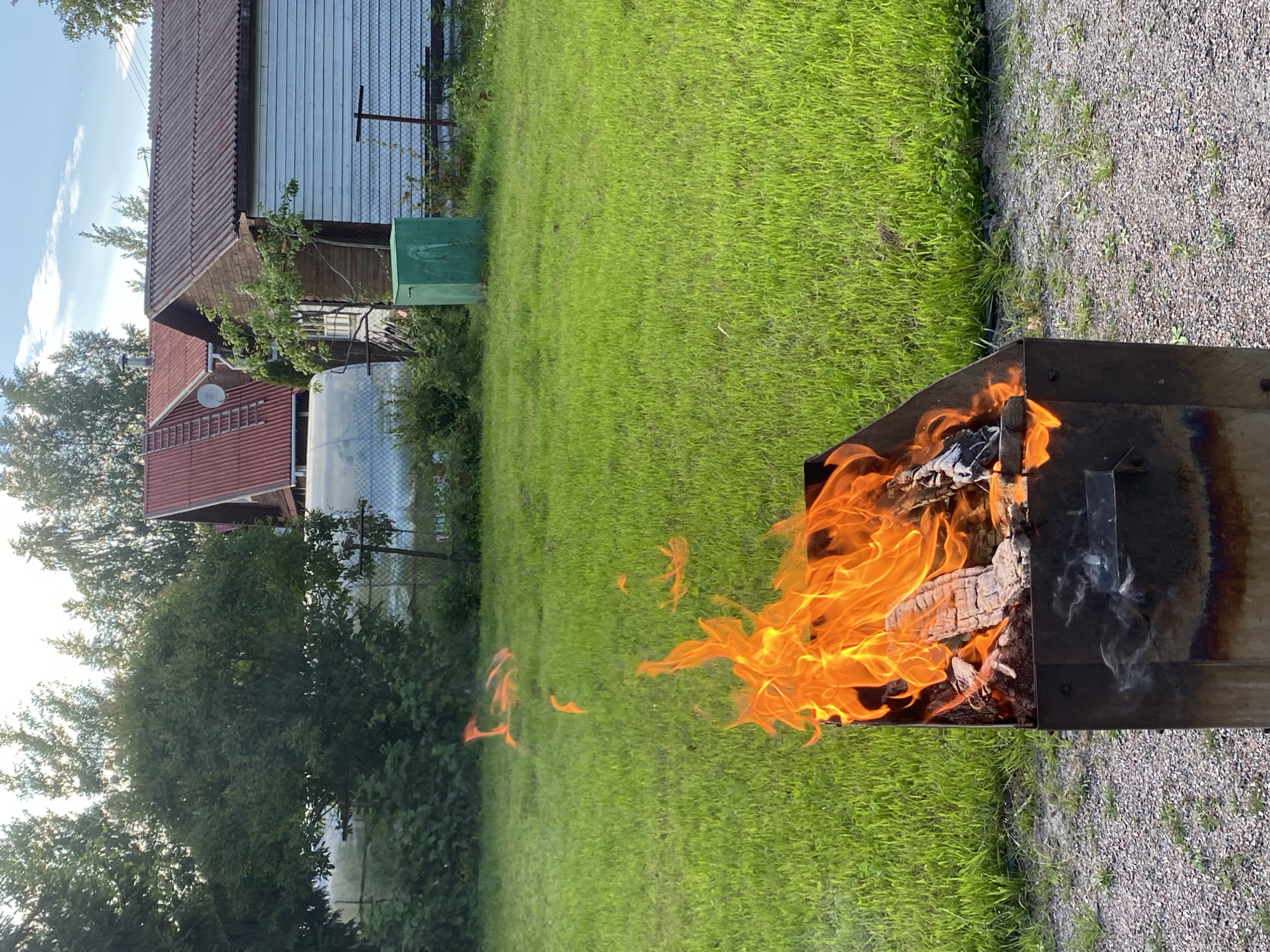 In Leipyasuo, Russia, a metal brazier stands on a gravel surface, with a vibrant fire burning inside. The flames are predominantly orange and yellow, consuming pieces of wood. The brazier is positioned in front of a lush green lawn, which extends to the background. Beyond the lawn, several houses with red tiled roofs are visible, nestled among trees. To the right of the houses, a green metal container is placed. The sky above is blue with scattered white clouds, suggesting fair weather. The scene captures an outdoor setting with a fire as the central focus.