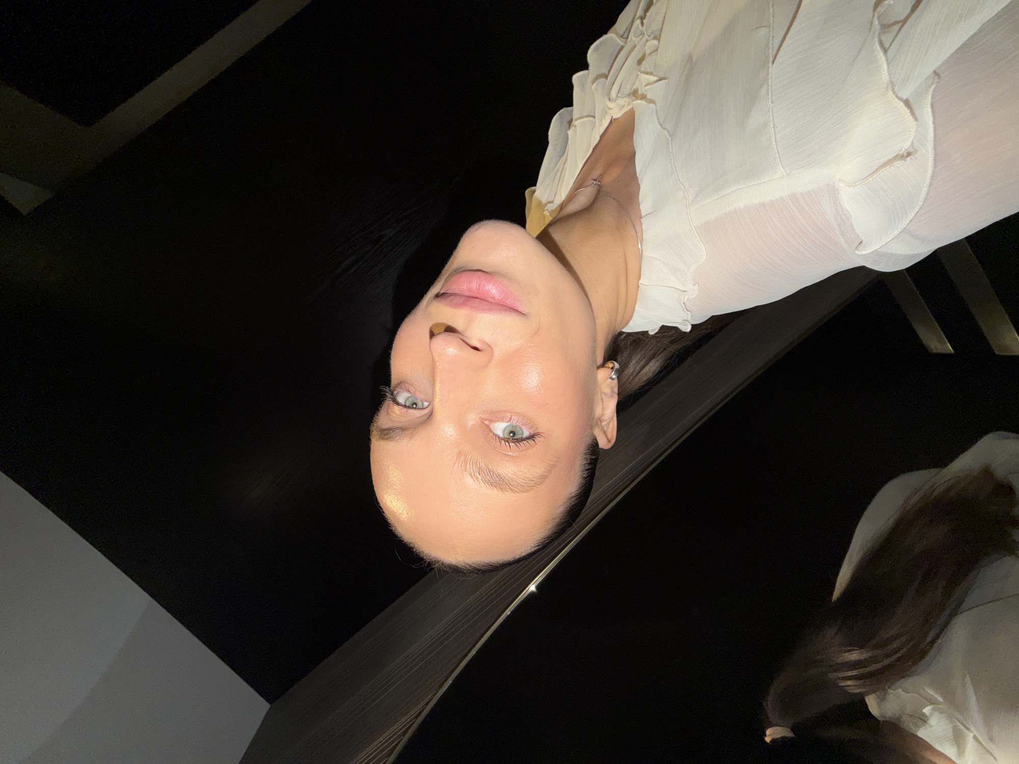 The image shows a close-up, slightly tilted shot of a young woman with light skin and blue eyes. She is wearing a light-colored, ruffled top and a delicate silver necklace. Her hair is long and dark. The background is dark and out of focus, with diagonal lines suggesting an architectural element. The scene appears to be indoors, possibly in a dimly lit room with reflective surfaces. There are no discernible elements that would indicate a specific location in Budva, Montenegro.