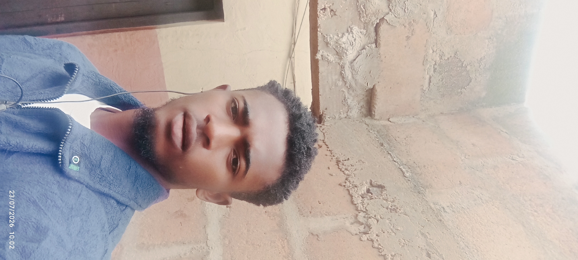 A medium shot captures a young Black male with dark, curly hair and a short beard from the chest up, standing in front of a textured, light brown brick wall. He is wearing a dark blue fleece jacket over a white t-shirt. Earbuds are visible, with the wire extending down his jacket. The background wall shows signs of wear and tear, with some areas of discolored plaster. To the left of the frame, a portion of a light brown structure, possibly a doorway or wall, is visible. A date and time stamp, "23/07/2026 10:02", is present in the lower right corner of the image. The lighting appears to be natural, casting a soft glow on the subject. The scene is consistent with an outdoor urban environment in Nsukka, Nigeria, likely in a residential or less-maintained area, given the weathered brickwork and simple structures.