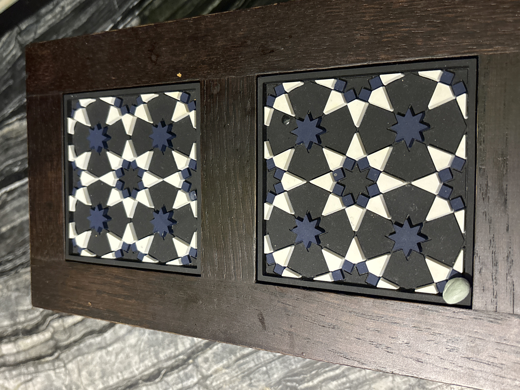 The image shows a close-up, overhead view of a wooden box with two decorative geometric inlays. The inlays are dark gray with a repeating pattern of white triangles and dark blue eight-pointed stars. The box is resting on a textured surface that appears to be fabric, possibly a blanket or tablecloth, with a marbled pattern of gray and white. A small, oval, light green object, possibly a stone or bead, is visible in the bottom left corner of the frame, resting on the wooden edge of the box. The scene appears to be indoors, and no specific location in Dubai, United Arab Emirates, can be determined from the image.