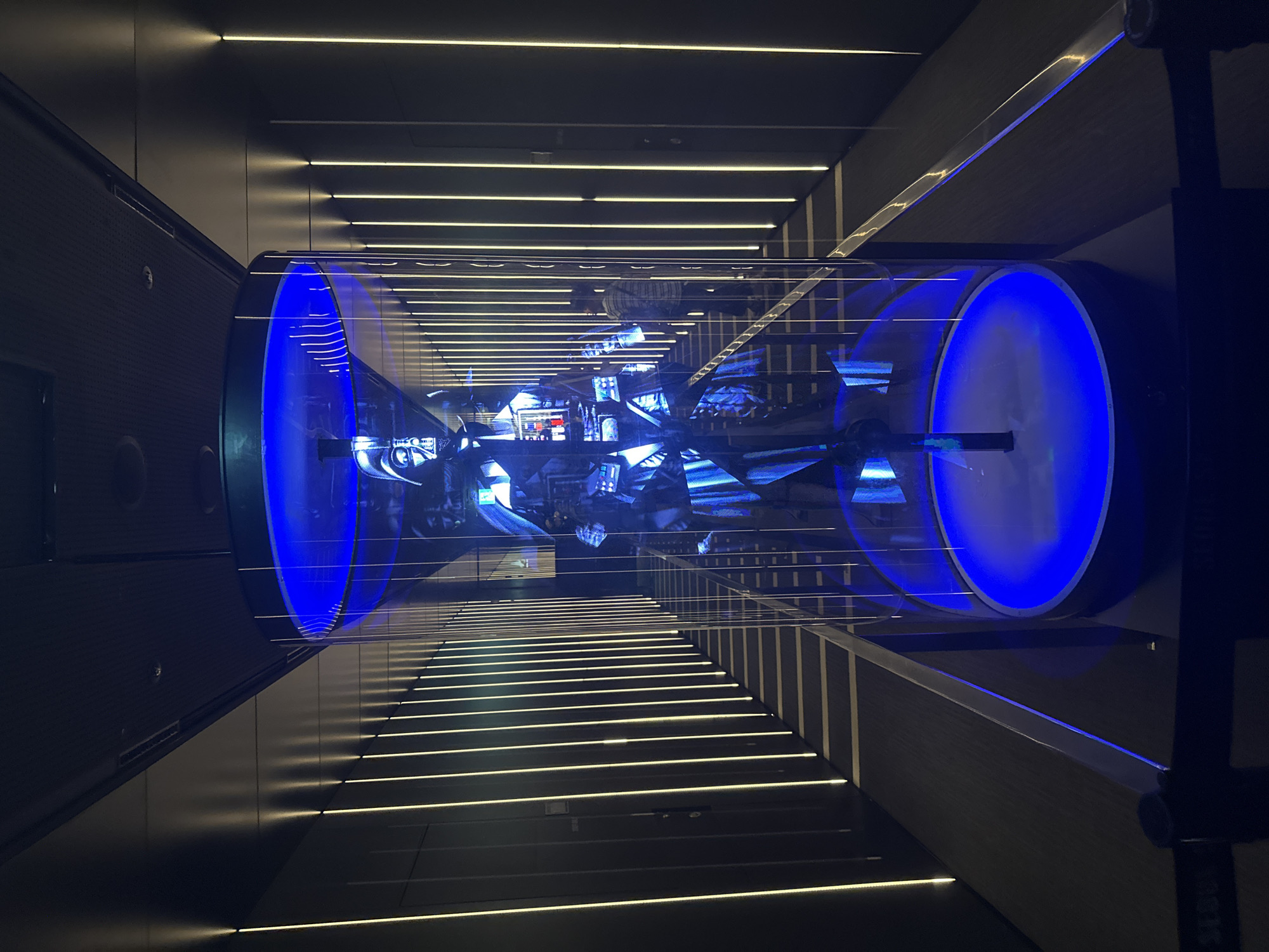 The image depicts an art installation within a building in Seoul, South Korea. The installation consists of a transparent cylindrical structure with blue illuminated circular ends. The cylinder contains a miniature cityscape. The surrounding architecture features parallel linear lighting embedded in dark surfaces, giving a perspective towards the installation.