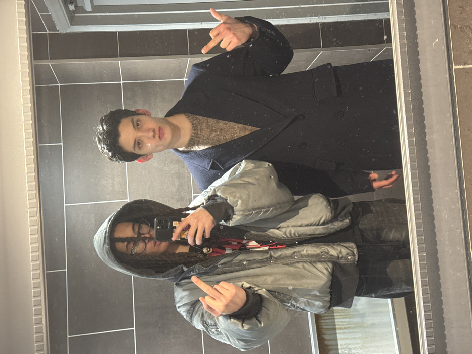 Two people are reflected in a mirror, rotated. One person wears a dark coat over a brown sweater and holds up their hand with two fingers extended. The other wears a hooded jacket, glasses, and dreadlocks, holding up their middle finger. Both appear to be posing for a selfie in front of a tiled wall. The overall image seems casual and perhaps playful, despite the offensive gesture.