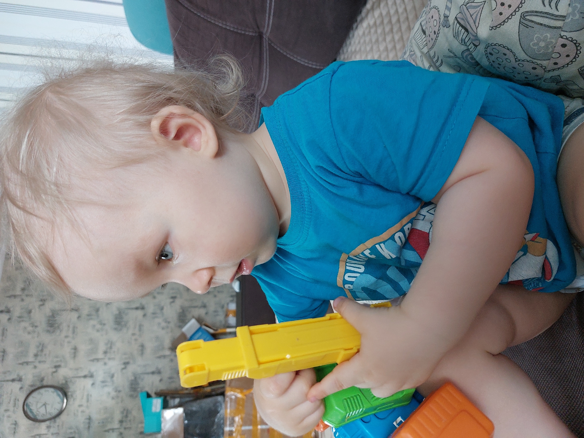 A young child with blond hair and fair skin is seated, wearing a blue t-shirt. The child is holding a multi-colored plastic toy gun, with the dominant colors being yellow, green, blue, and orange. The child is looking down at the toy. The child is situated on a couch with patterned cushions and a darker brown upholstered section. The background includes a wall with a patterned wallpaper and a clock. Based on the request, the location is identified as Chaikovsky, Russia, but no specific landmarks or indicators in the image confirm this.