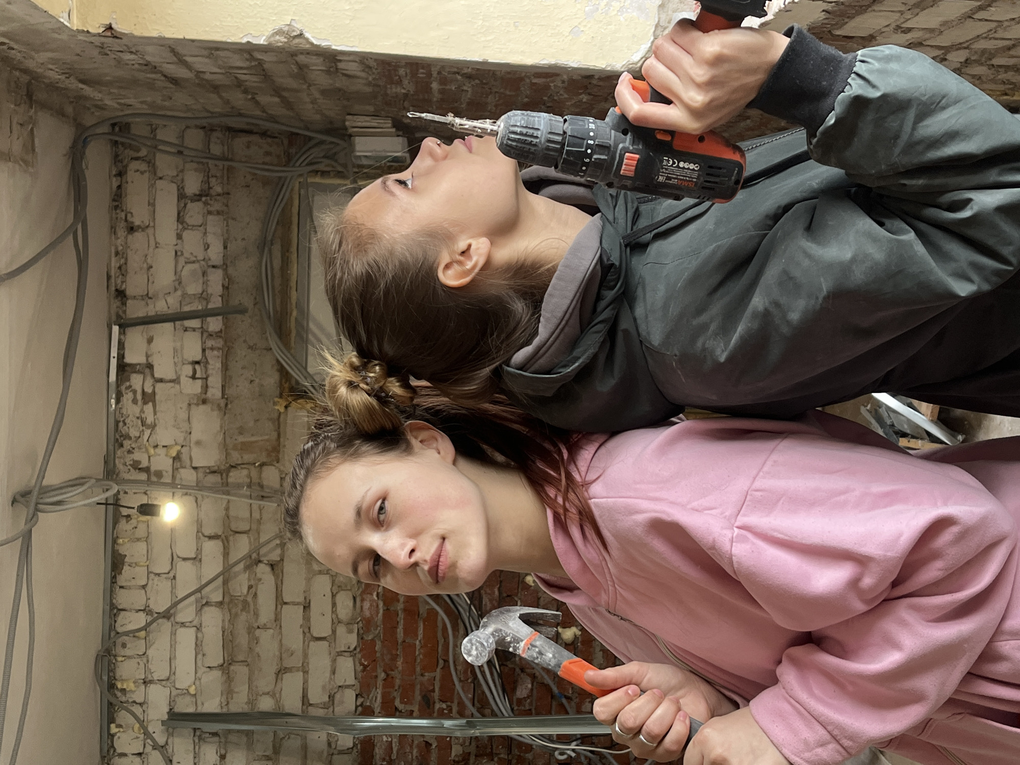 Two young women are pictured in an interior setting with exposed brick and wiring, presumably undergoing renovation. The woman in the pink hoodie holds a hammer, while the woman above her holds a power drill. The environment appears to be in a state of construction or repair. There is no information indicating it is located in Фоки, Russia.