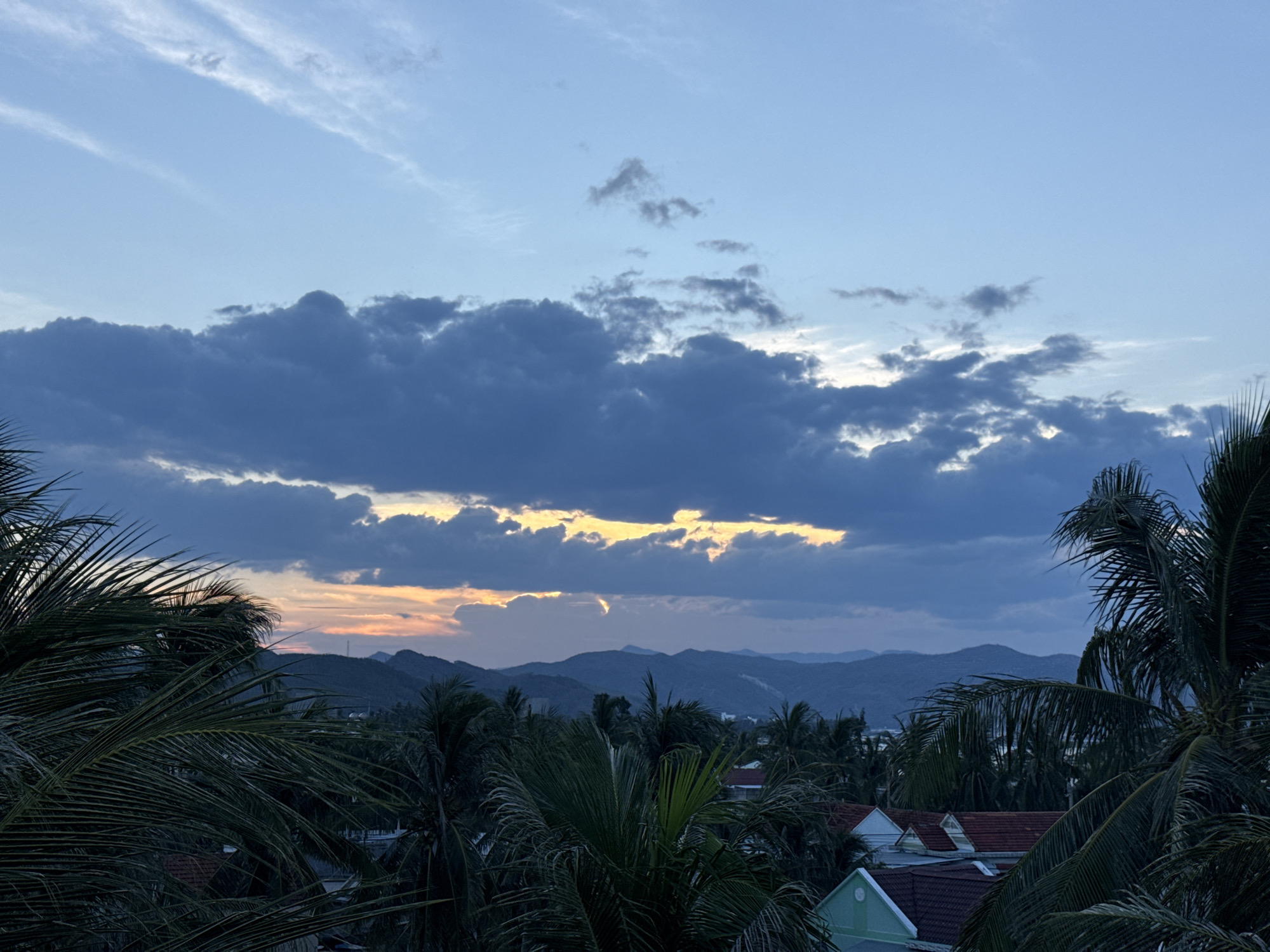 The image depicts a coastal scene at dusk, with a sky filled with dramatic clouds that allow a sliver of orange and yellow sunset light to peek through. In the foreground, lush green palm fronds frame the view, obscuring parts of the landscape below. Beyond the palms, a dense canopy of trees stretches across the middle ground, interspersed with the red-tiled roofs of houses, some with white walls and green accents. In the distance, rolling, dark blue mountains create a layered silhouette against the fading light. The overall impression is one of a tropical evening, with the natural beauty of the landscape dominating the scene.