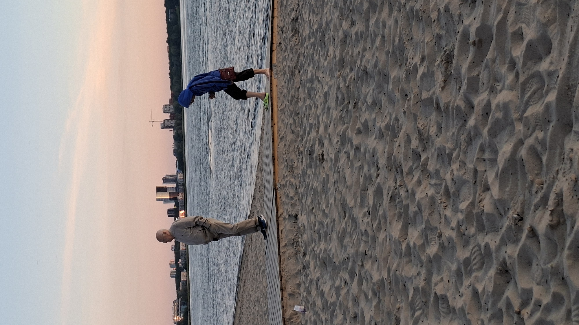 A man and a child are on a beach near the water at sunset. The man is older, wearing a light-colored jacket and pants, and is standing still, looking out at the water. The child is wearing a blue hoodie and dark pants, and appears to be walking or playing near the water's edge. The beach is sandy with wooden boardwalks. In the background, a cityscape with buildings and cranes can be seen across the water. The sky is a soft pink and orange, indicating the time of day is sunset. The image is taken from a low angle, emphasizing the sand in the foreground. The location is the Pirita beach in Tallinn, Estonia.