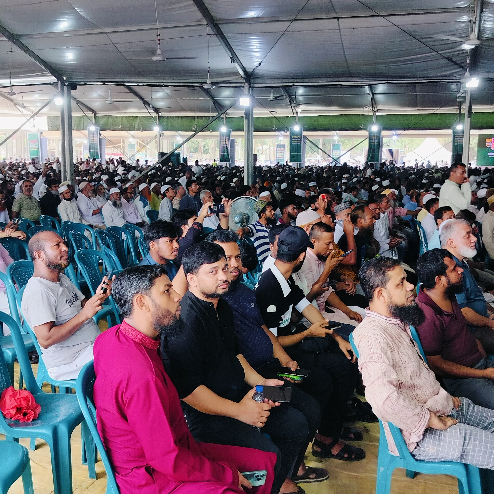A large gathering of men fills a tent-like structure in Dhaka, Bangladesh. The men are seated in blue plastic chairs, facing in the same direction. Some are using mobile phones, while others are looking ahead. The men are dressed in various styles of clothing. Overhead lighting hangs from the tent ceiling. Banners line the back of the tent. The crowd extends into the distance.