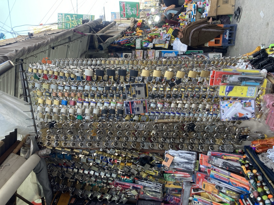 The image portrays an outdoor market stall in Quetta City Tehsil, Pakistan. The foreground is dominated by a dense display of hundreds of padlocks of varying sizes, shapes, and materials, predominantly silver-toned, brass, and black, with some featuring colored elements (blue, red, green). These padlocks are individually hung from metal rods in numerous vertical rows. Below the padlocks, shelves display packaged consumer hardware items, including multi-packs of pliers, a white hot glue gun, and a soldering iron kit.
To the right, a male individual is seated on a blue plastic crate, draped in a patterned brown shawl that covers his body. He wears a dark cap and looks towards the left. In the mid-ground and background, further market activity is visible, with other stalls displaying various goods and several individuals present, some standing or walking. Overhead, elements of market infrastructure are visible, including awnings, exposed wiring, and two prominent green signs with white Urdu script on a building facade. The environment is an active street market under an overcast daytime sky. No direct interactions between individuals are discernable.