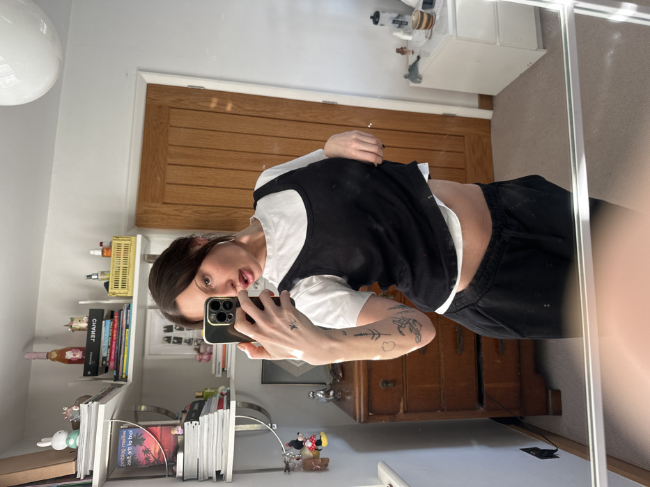 A female individual is depicted taking a mirror selfie within a domestic interior, located in the United Kingdom. She stands facing the mirror, holding a dark-colored smartphone in her right hand to capture the image. Her dark hair is pulled back, and she wears a white short-sleeved t-shirt beneath a black scoop-neck tank top, paired with black drawstring sweatpants. Her left hand is positioned at her waist, subtly lifting the hem of the black tank top, revealing a segment of her midriff and the underlying white t-shirt. Visible tattoos include a star on her left wrist and botanical designs on her left forearm.
The background displays an organized room with off-white walls and light-colored carpet. To the left, two white wall-mounted shelves hold stacks of books, a pink champagne bottle, a light green rabbit figurine, and miscellaneous decorative objects, including Minnie Mouse keychains. Below the shelves, a wooden dresser with four drawers is present. To the right, a light brown wooden paneled door is visible, adjacent to a white two-drawer bedside table topped with various items. A white spherical ceiling light fixture is partially observable in the upper right quadrant. No visual cues within the image definitively identify the specific location as Wealden, beyond its classification as a domestic interior in the UK.