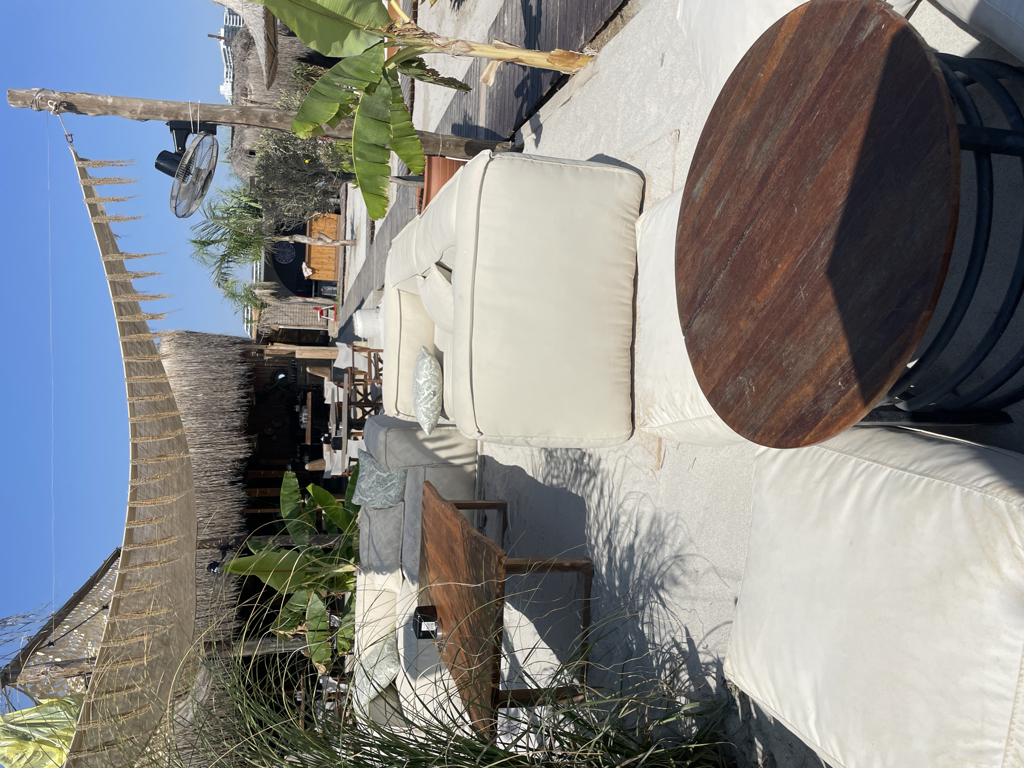 The image depicts an outdoor seating area at a beachside establishment in Trikomo, Northern Cyprus. The scene is set on a sandy beach with a clear blue sky overhead. In the foreground, a round wooden coffee table with a dark metal base rests on a large, white floor cushion. Behind this, a large, L-shaped white sofa with light blue patterned cushions is prominently featured. To the left of the sofa, a rectangular wooden coffee table with a live-edge top sits on the sand.
The background reveals a rustic, open-air structure with thatched roofs and canvas awnings providing shade. Tropical plants, including banana trees and palm trees, are scattered throughout the area. A large industrial fan is mounted on a wooden post to the right, indicating an effort to combat the heat. Wooden boardwalks are visible, suggesting pathways through the sand. In the distance, modern buildings are partially visible, contrasting with the natural and rustic elements of the beach club. No people are present in the image. The overall atmosphere is one of a relaxed, sunny, beachside resort.