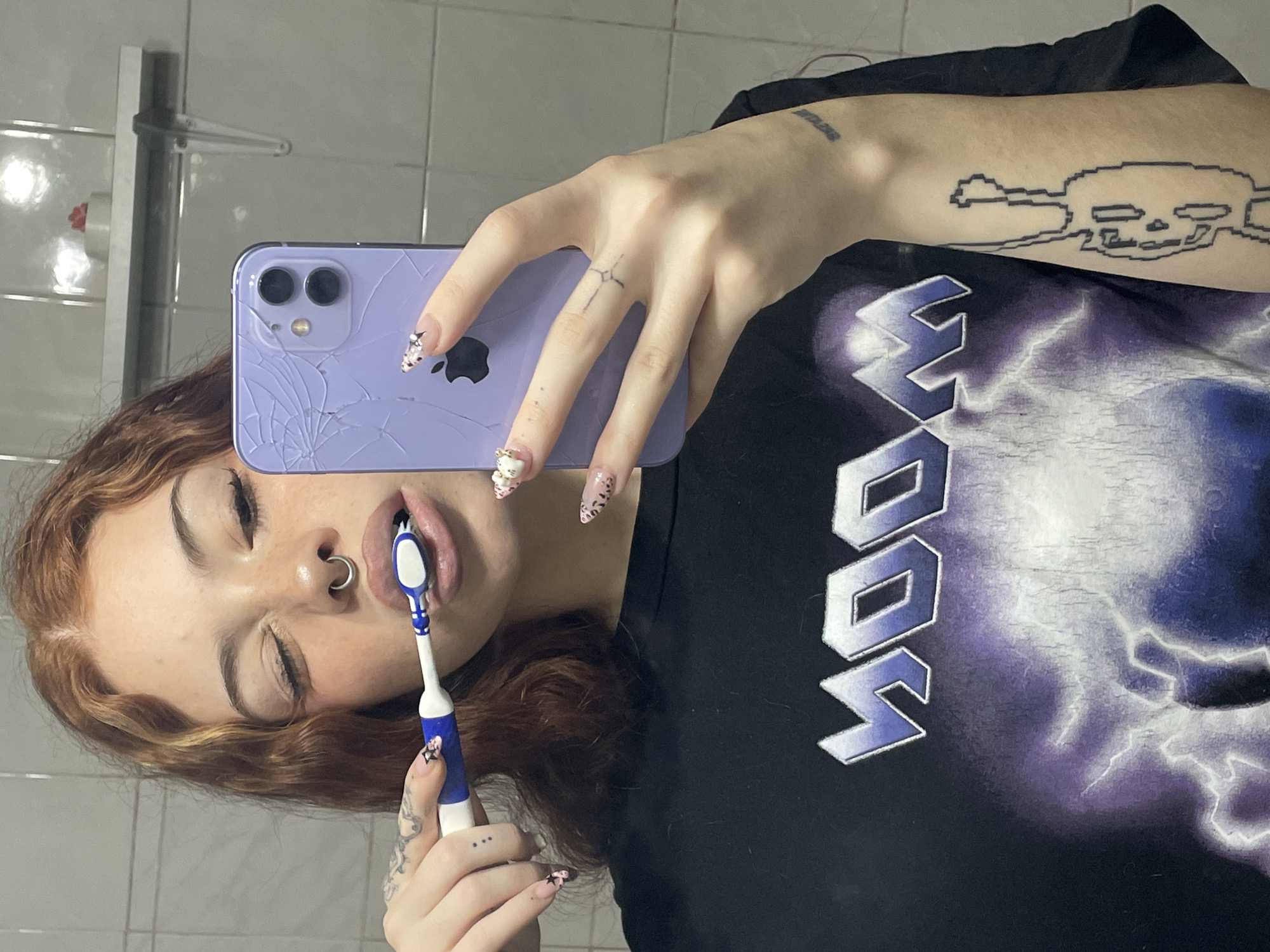 A young woman is taking a selfie while brushing her teeth in a bathroom. She is fair-skinned with reddish-brown hair and multiple tattoos on her fingers and arms. She has a nose piercing. She is holding a purple iPhone with a cracked screen to take the picture. Her nails are decorated with designs. She is wearing a black t-shirt with a graphic print. The bathroom has white tiled walls and a chrome towel rack. It is impossible to determine if the location is Buenos Aires, Argentina from the image alone.