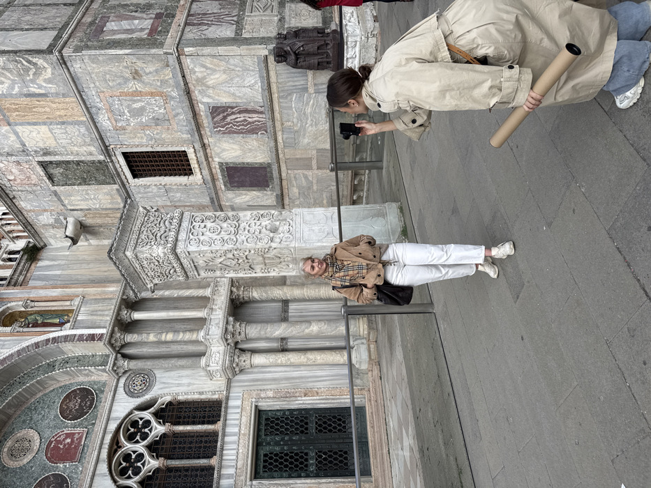 The scene depicts two adult females on a grey paved walkway in front of St. Mark's Basilica in Venice, Italy. The background is dominated by the Basilica's ornate facade, characterized by multiple arches, marble columns with intricate capitals, varied marble cladding, decorative mosaics, and grid-patterned windows. Part of the Porphyry Group sculpture (Portrait of the Four Tetrarchs) is visible on the far right. A low, horizontal barrier separates the individuals from the immediate building structure.
In the left-midground, a blonde-haired female, wearing a light brown cropped jacket, a plaid scarf, white high-waisted trousers, and white athletic shoes, stands facing approximately 45 degrees to her right, with hands in her pockets and a subtle smile. A dark shoulder bag hangs from her left shoulder.
In the right foreground, a dark-haired female, wearing a long beige trench coat, light blue jeans, and white athletic shoes, is crouched. She holds a black digital camera with both hands, aiming it directly towards the first female. Her left hand also holds a brown cardboard tube, pointed downwards.