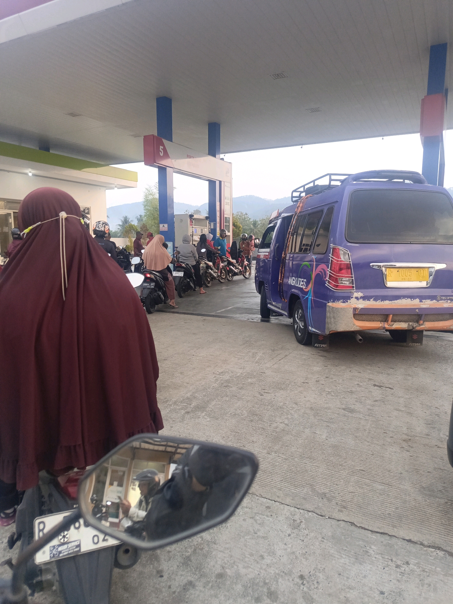 A line of motorcycles and a purple van are at a gas station under a covered canopy in Pegalongan, Indonesia. A person wearing a dark maroon hijab is in the foreground, facing away from the camera, seated on a motorcycle. Behind them, several other motorcycles and their riders are waiting in line. A purple van with the word "ANGKUDES" and colorful graphics is parked to the right, facing the viewer. The gas station has blue support poles and signs with the number "5" and "PERTAMINA". The ground is paved with concrete, and in the background, hills can be seen. The scene appears to be during daylight hours.