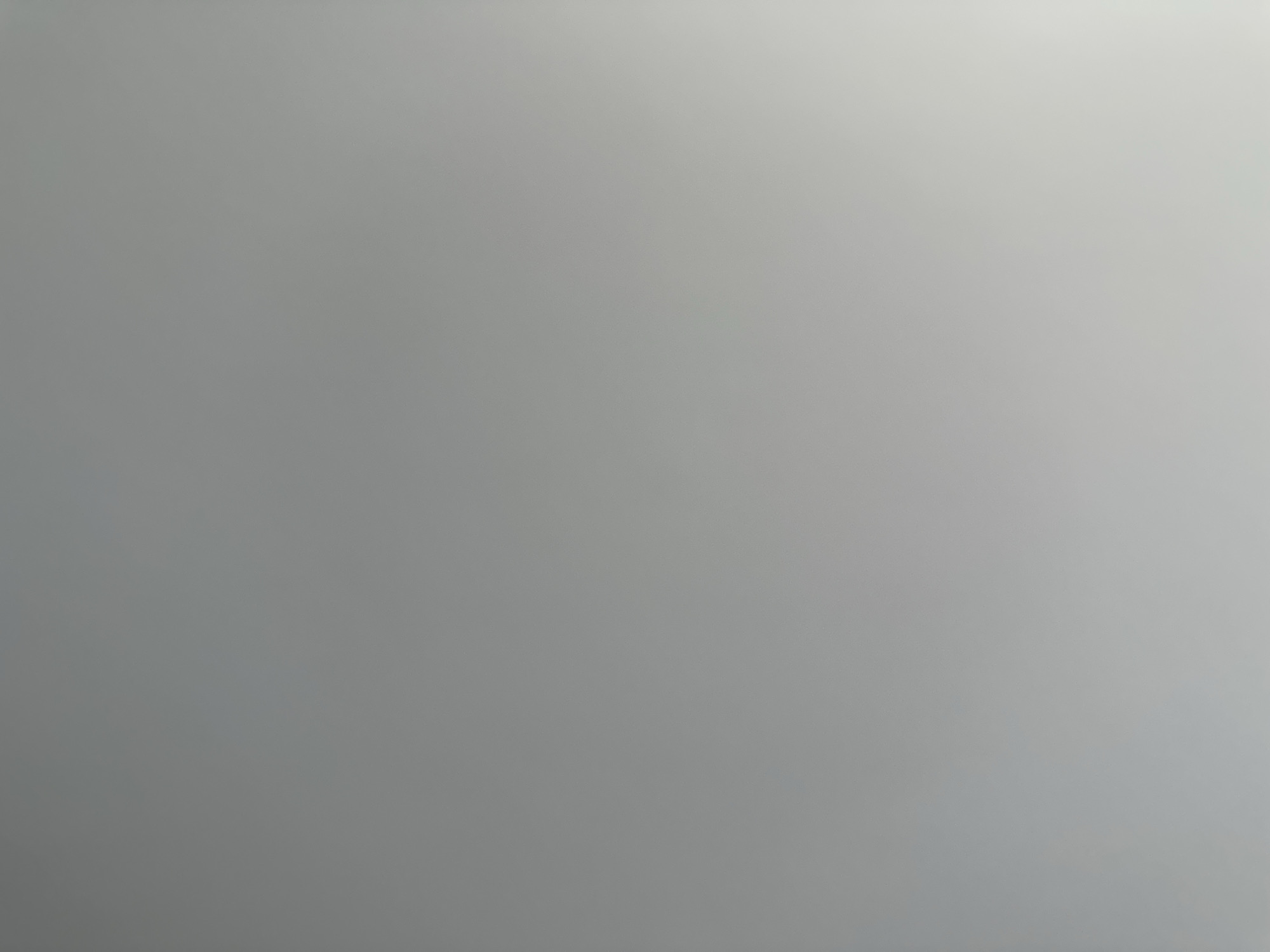 The image is a close-up, high-angle shot of a gray, textured wall. The texture appears to be a smooth, slightly uneven surface, possibly plaster or painted concrete. There are subtle variations in shading across the wall, with a lighter area towards the bottom right corner, suggesting a light source from that direction. No objects, people, or discernible features are present in the image. The scene does not provide any visual cues that would allow for a specific location to be identified, including within the city of Belgrade, Serbia.